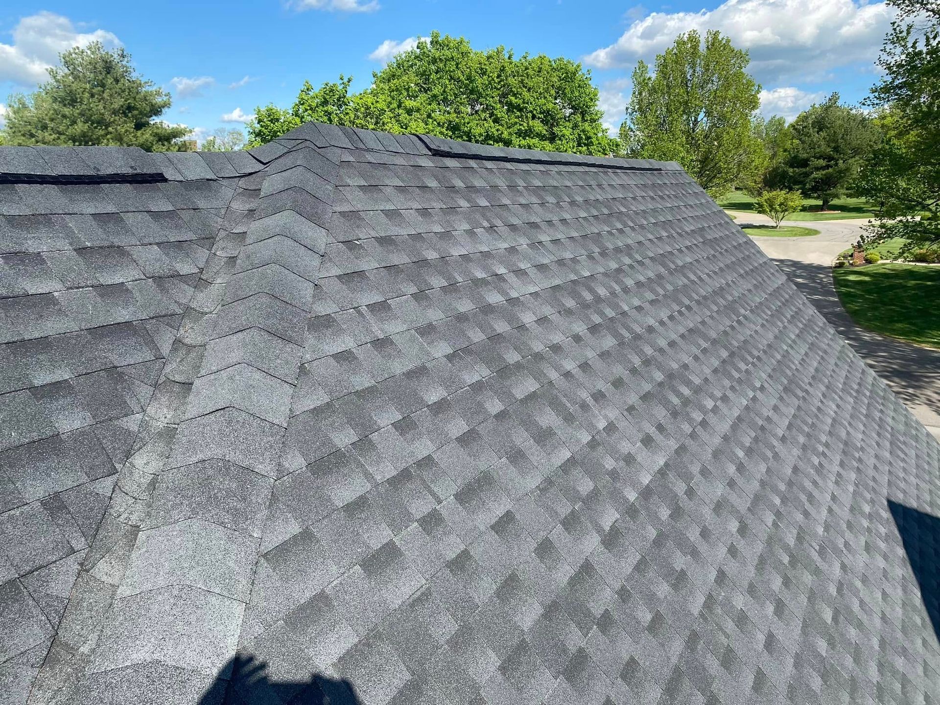 Gray shingled roof with a roof vent under a blue sky and green trees