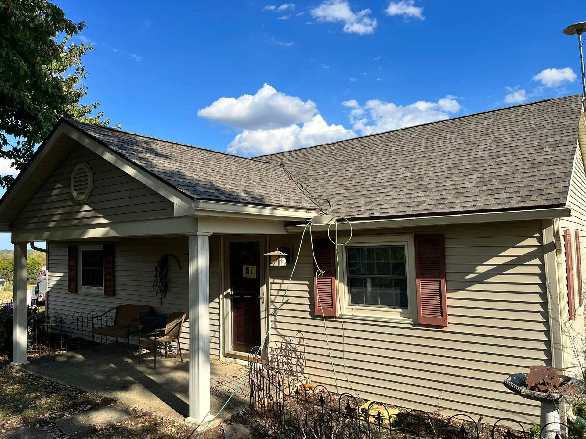 Single-story beige house with shingled roof, front porch, and red shutters under a blue sky.