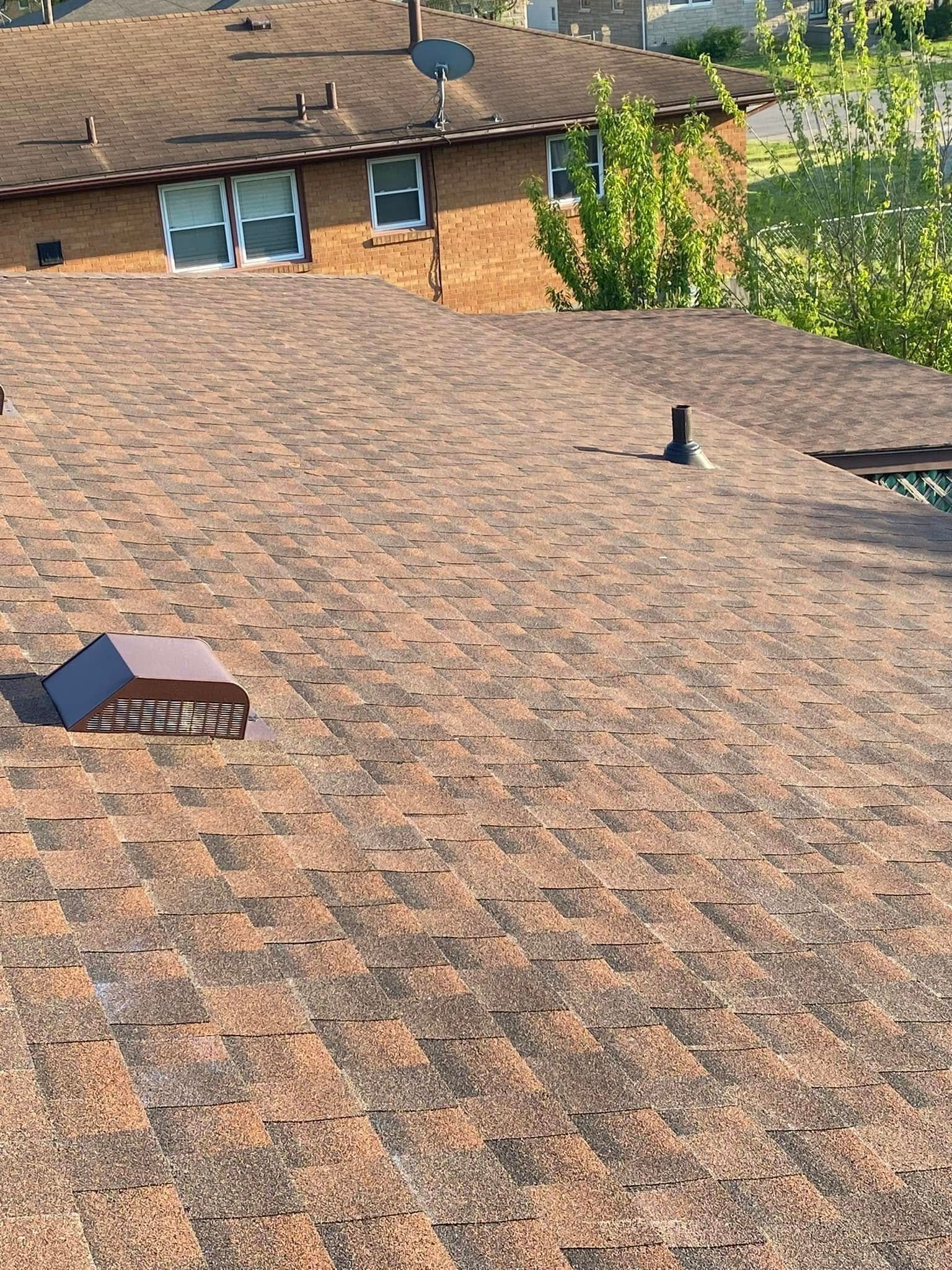 Roof with brown shingles, vents, and a brick house in the background near leafy trees