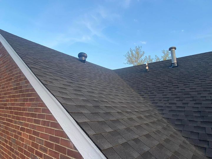 Shingled roof valley beside a red brick wall under a blue sky, with vents and a chimney pipe.