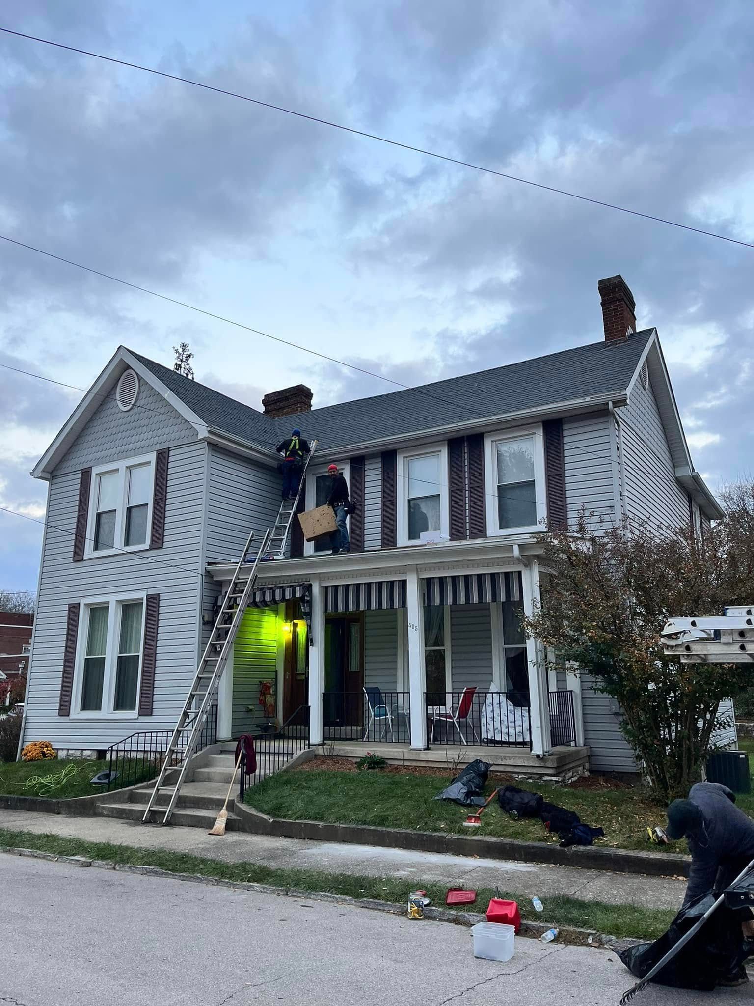 Two-story gray house with workers on a front porch and lawn under cloudy sky, lights glowing inside