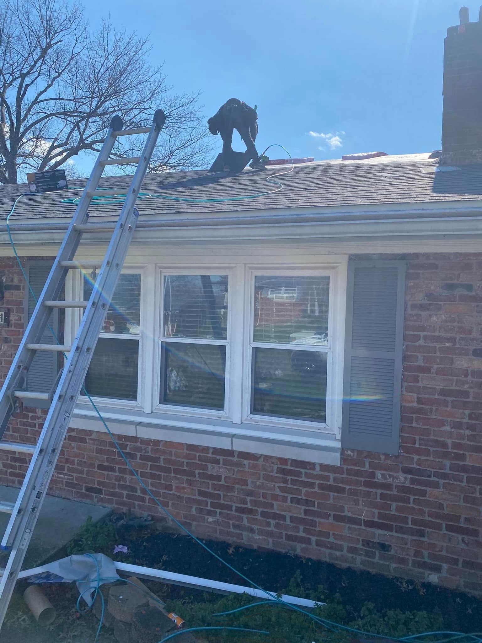 Worker on a roof beside a ladder, repairing shingles above a brick house with white windows.