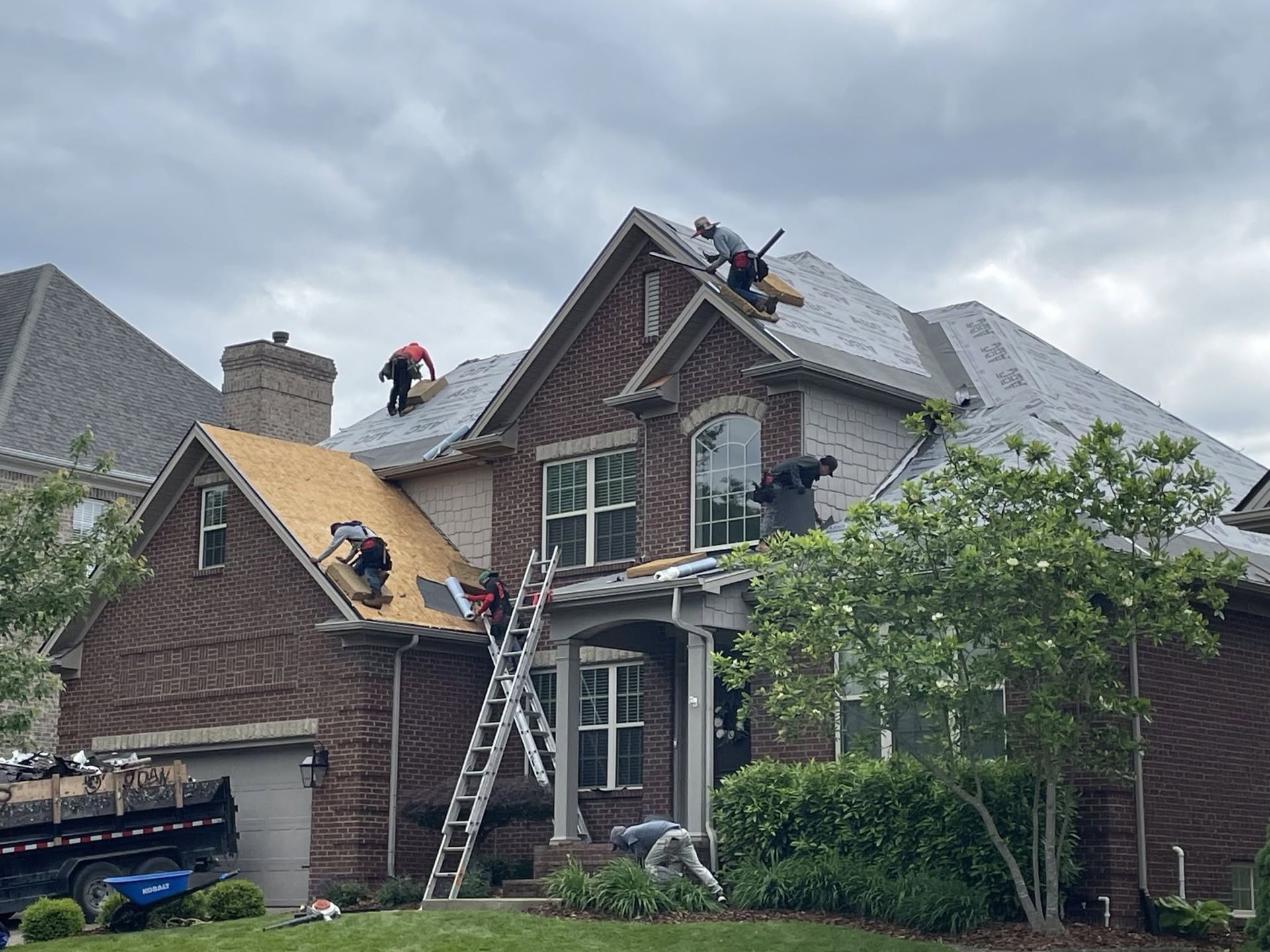 Workers repairing a two-story house roof with ladders under cloudy skies