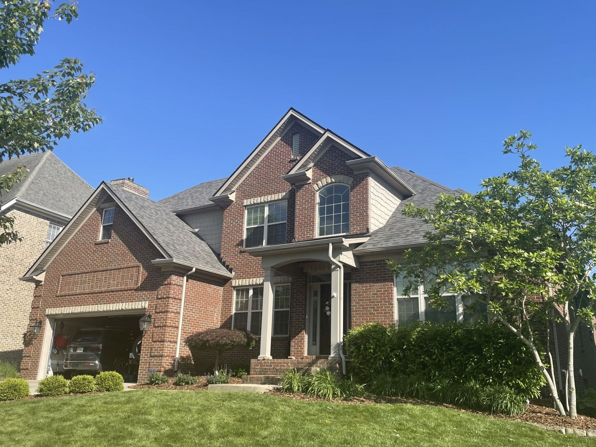Two-story brick suburban house with gabled roof, front porch, and manicured landscaping under a blue sky