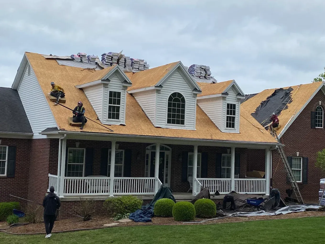 Damaged house roof with tarps and debris, workers on porch, cloudy sky