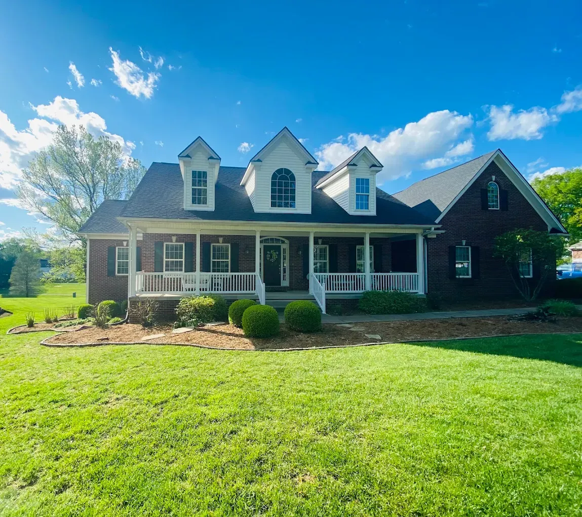 Sunny suburban house with stone accents, dormer windows, and a large green lawn under a blue sky