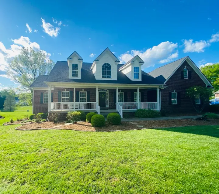 Brick house with white porch and dormer windows on a sunny day, set behind a green lawn.