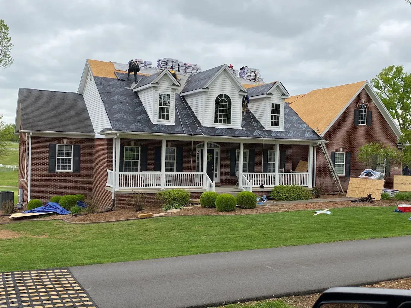 Large brick house with gray roof, white porch, and tan tarp-covered roof repair under cloudy sky