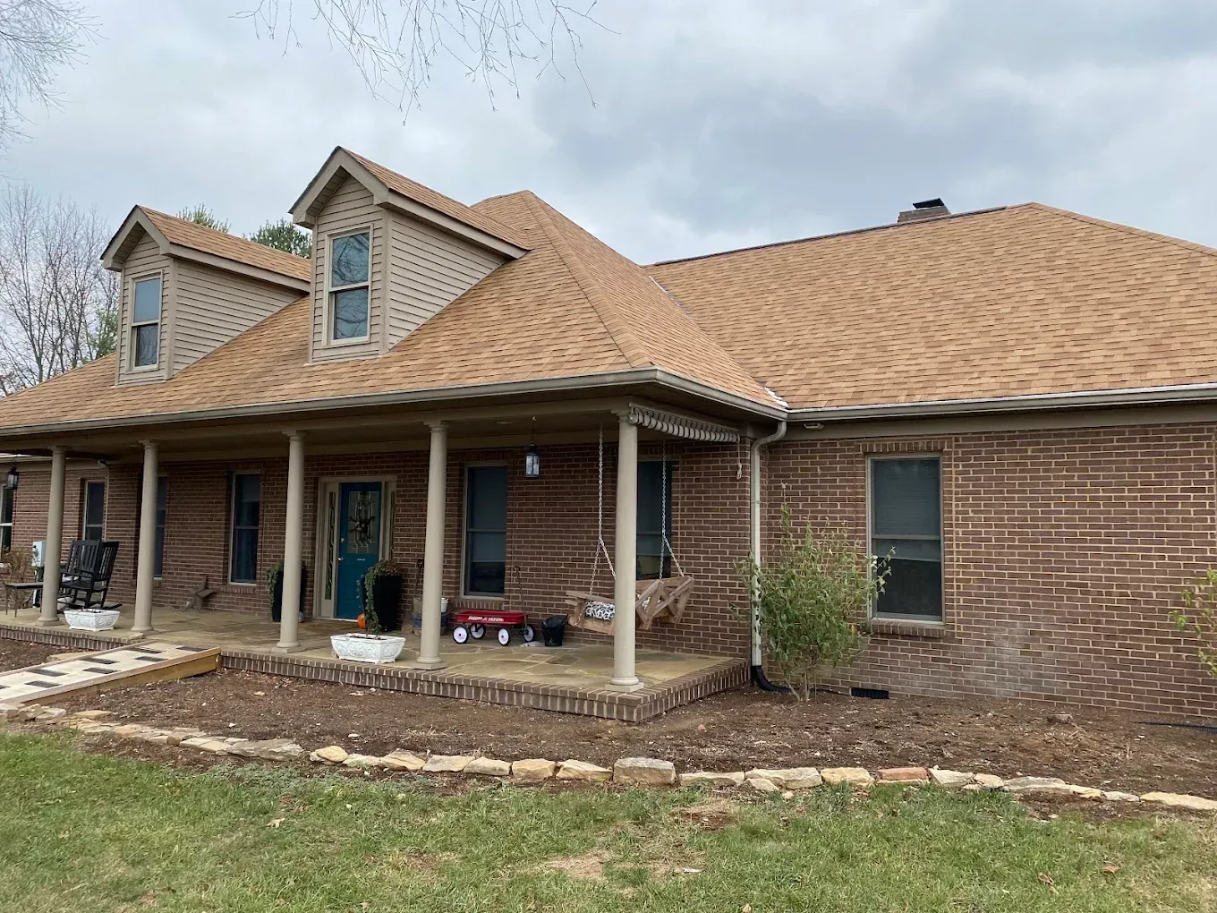 Single-story brick house with tan roof, front porch, and blue front door under a cloudy sky