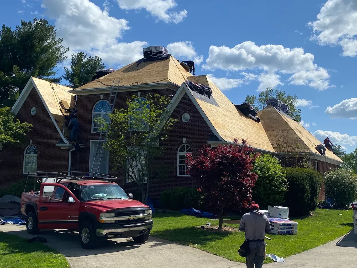 Red pickup truck parked by a large house with a tan roof under a bright blue sky with clouds
