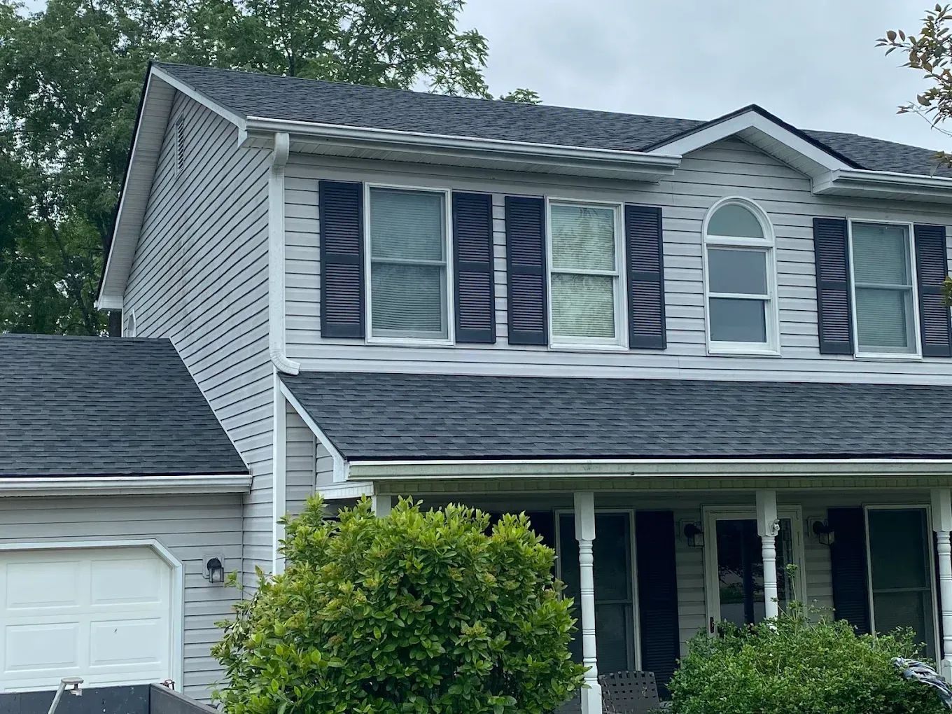 Two-story gray house with white trim, front porch, garage, and a bush in the yard