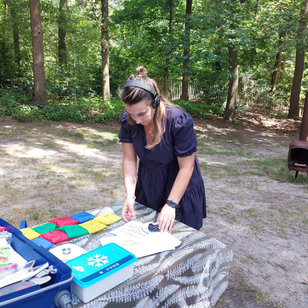 Vrouw die spullen schikt op een tafel met kleurrijke tafelkleden en een blauwe doos in een bosrijke omgeving.