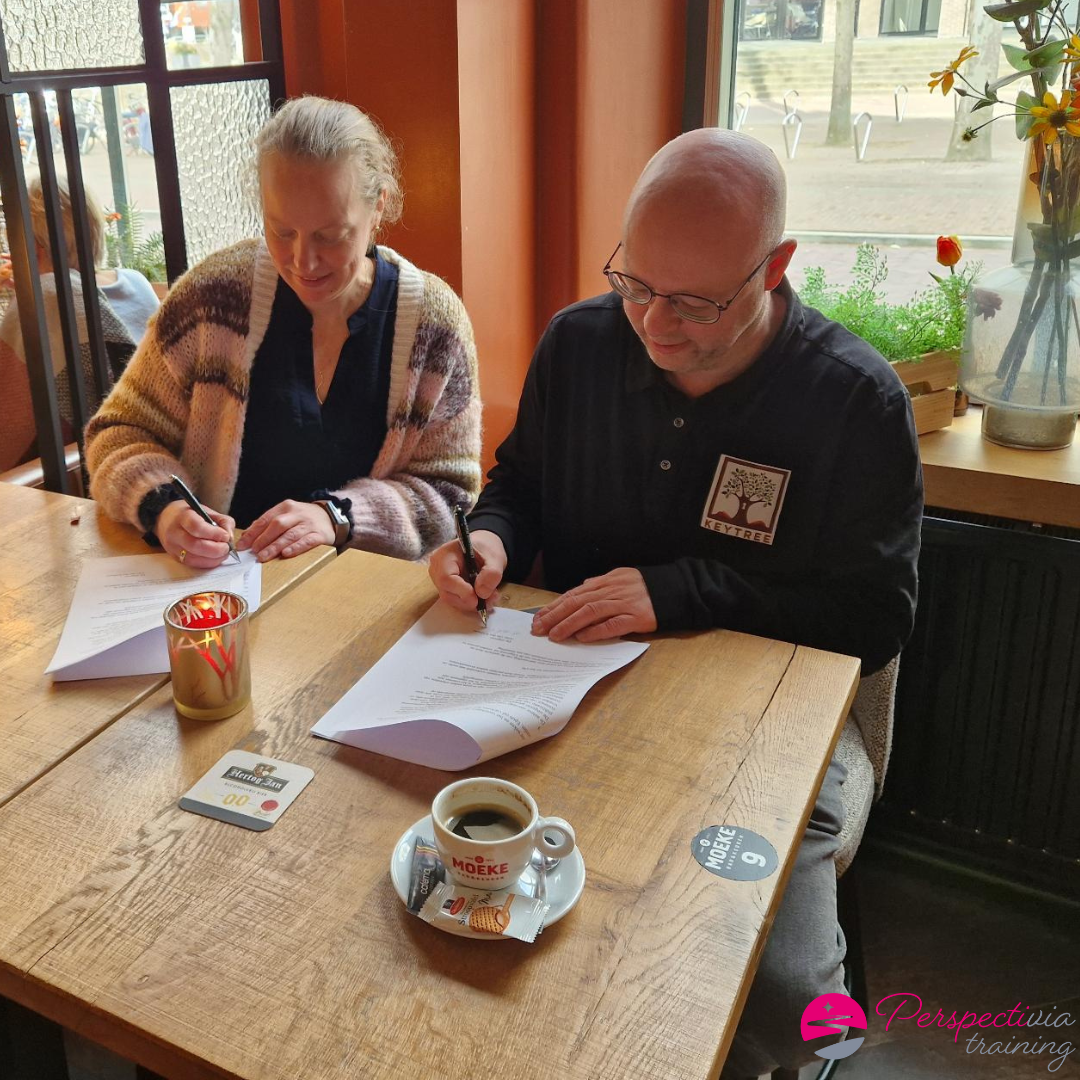Twee mensen zitten aan een houten cafétafel en schrijven met pennen op documenten. Tussen hen in staat een kop koffie op de tafel.