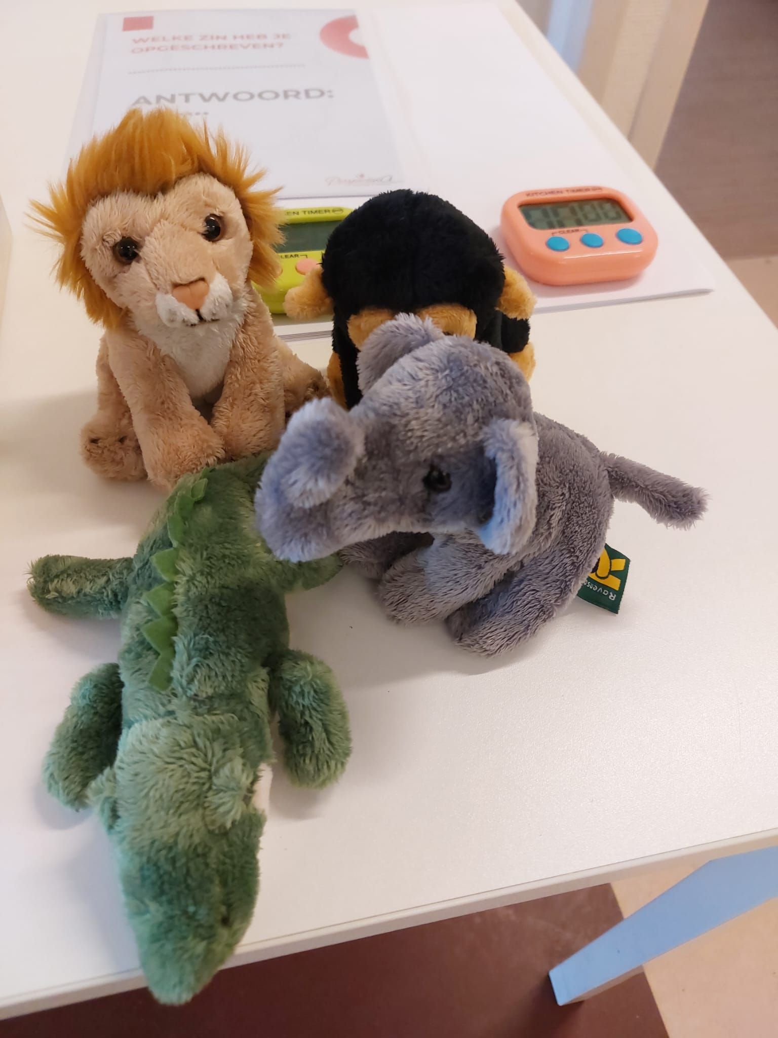Vier knuffeldieren – een leeuw, een aap, een olifant en een krokodil – staan op een witte tafel met een kleine timer.