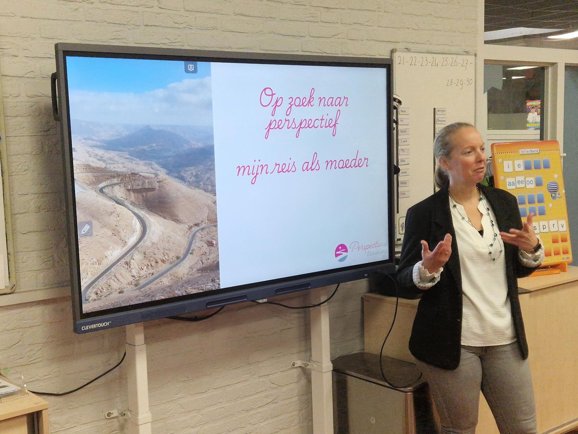 Vrouw spreekt naast een aan de muur gemonteerd scherm waarop een luchtfoto van de kustlijn en rode tekst te zien zijn.