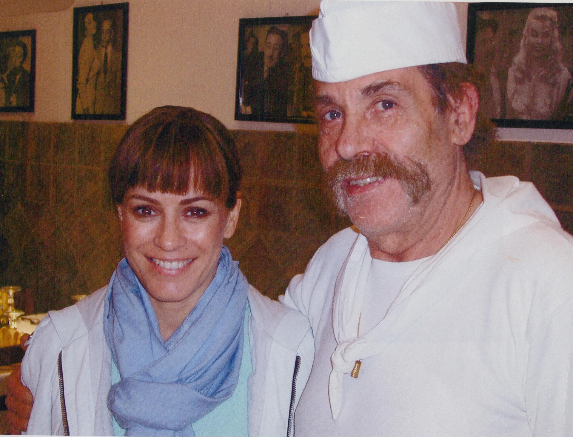 Una donna sorridente e uno chef con cappello e giacca bianchi in piedi in un ambiente interno con un quadro incorniciato appeso alla parete.