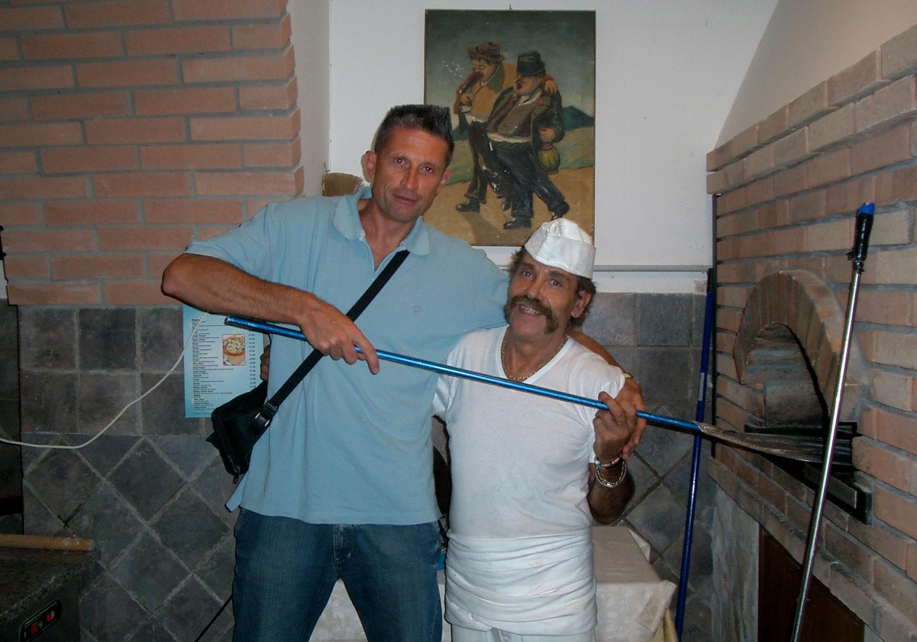 Due uomini posano in interni accanto a un camino in mattoni, tenendo in mano una lunga asta blu e sorridendo all'obiettivo.