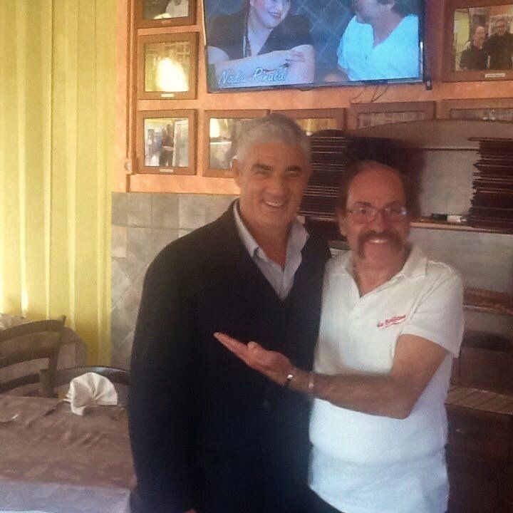 Due uomini sorridenti in posa in interni, uno con una giacca scura e l'altro con una polo bianca, con un televisore sullo sfondo.