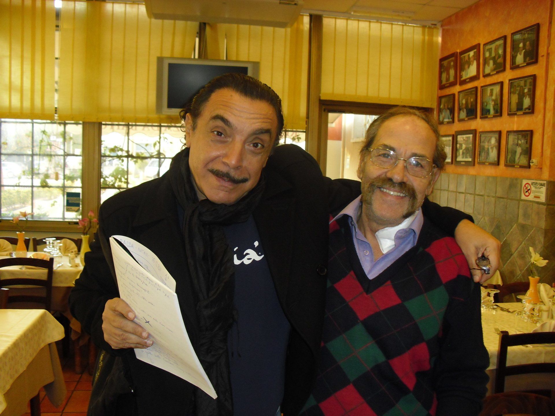 Due uomini sorridenti in un ristorante, uno con un giornale in mano e l'altro con un braccio intorno alle spalle.