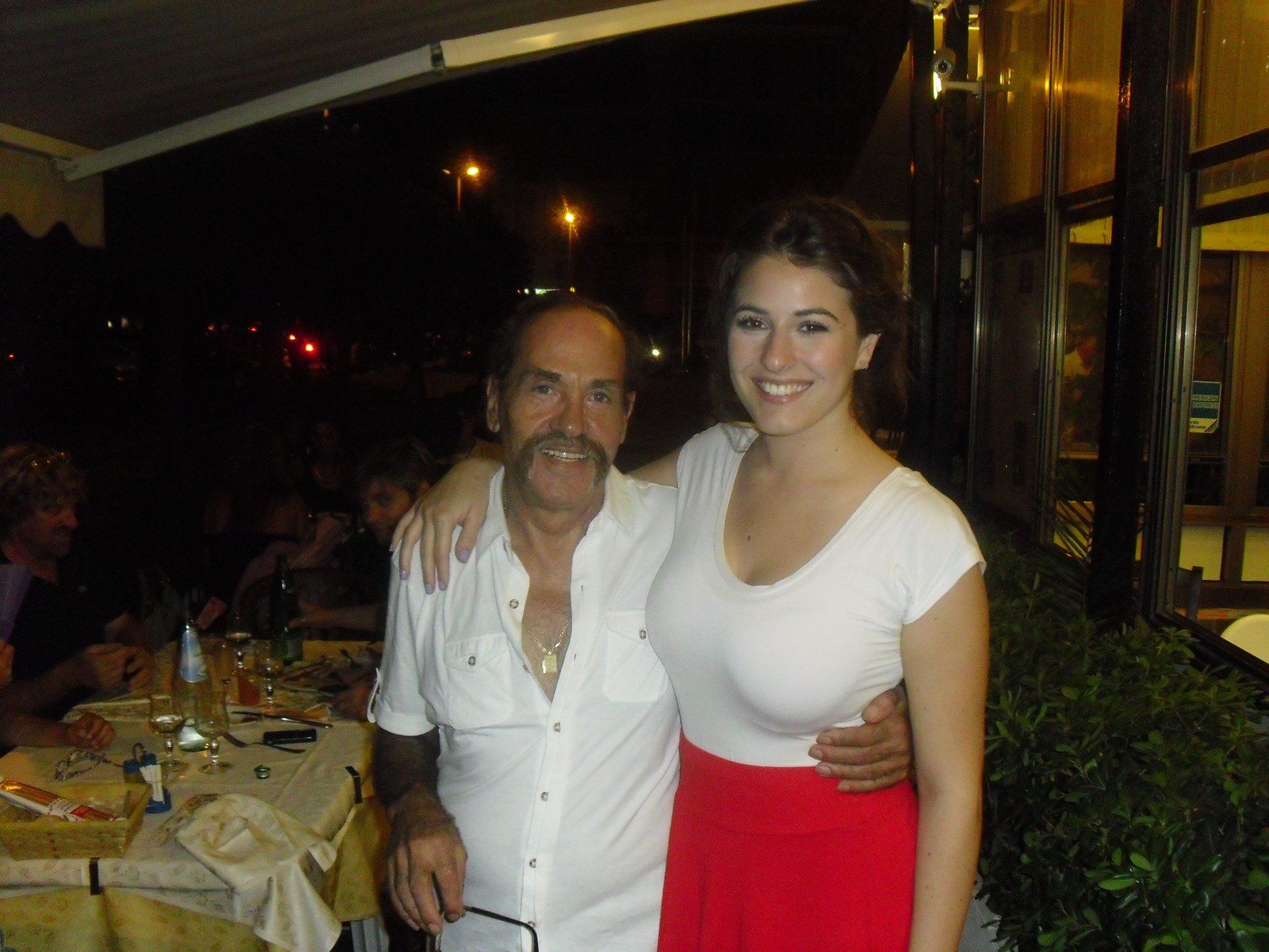 Due persone sorridenti in un ristorante all'aperto di sera, in piedi accanto a un tavolo con altri clienti sullo sfondo.