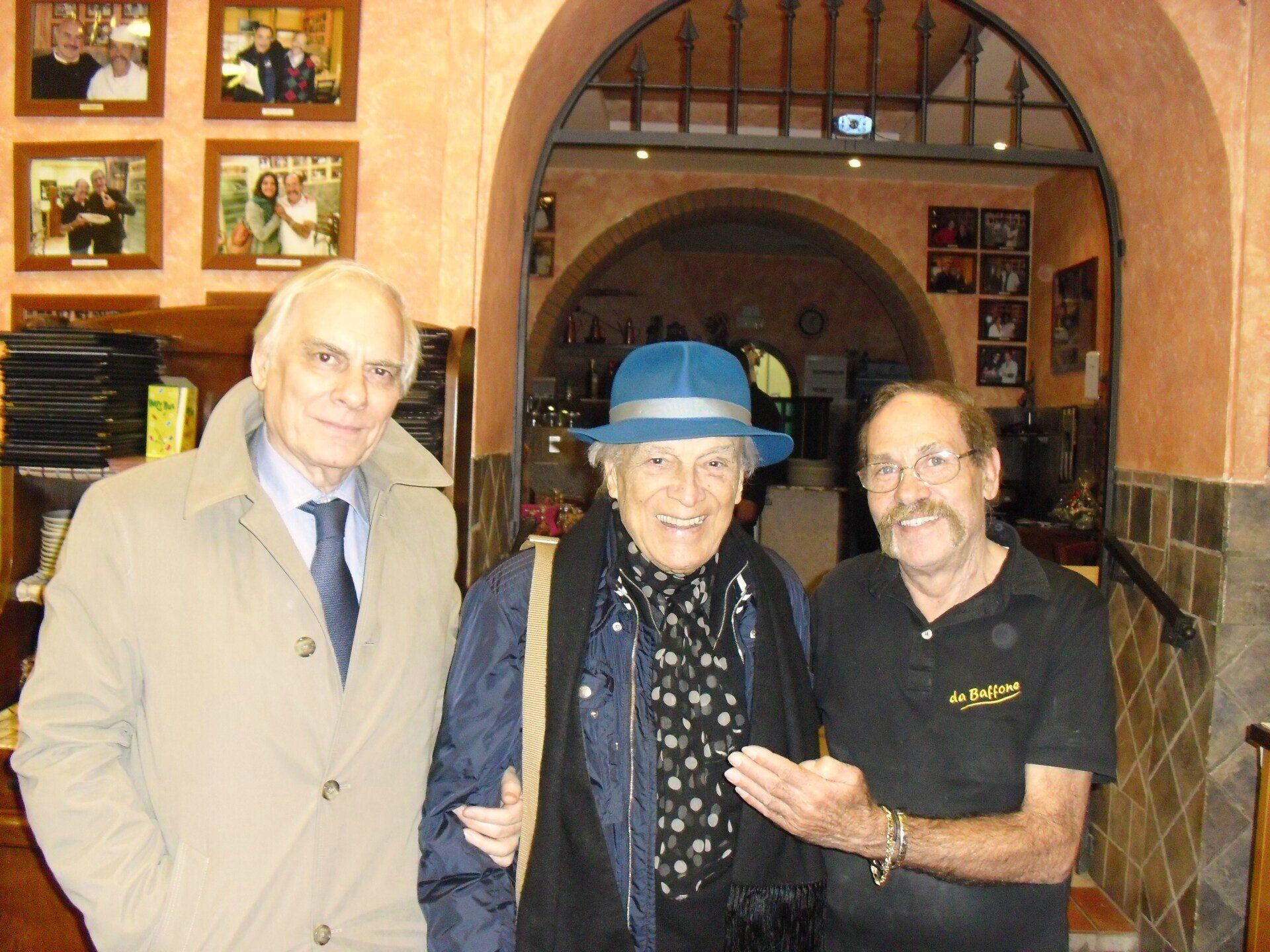 Tre persone in posa in interni, con foto incorniciate alle pareti e una porta ad arco alle loro spalle.