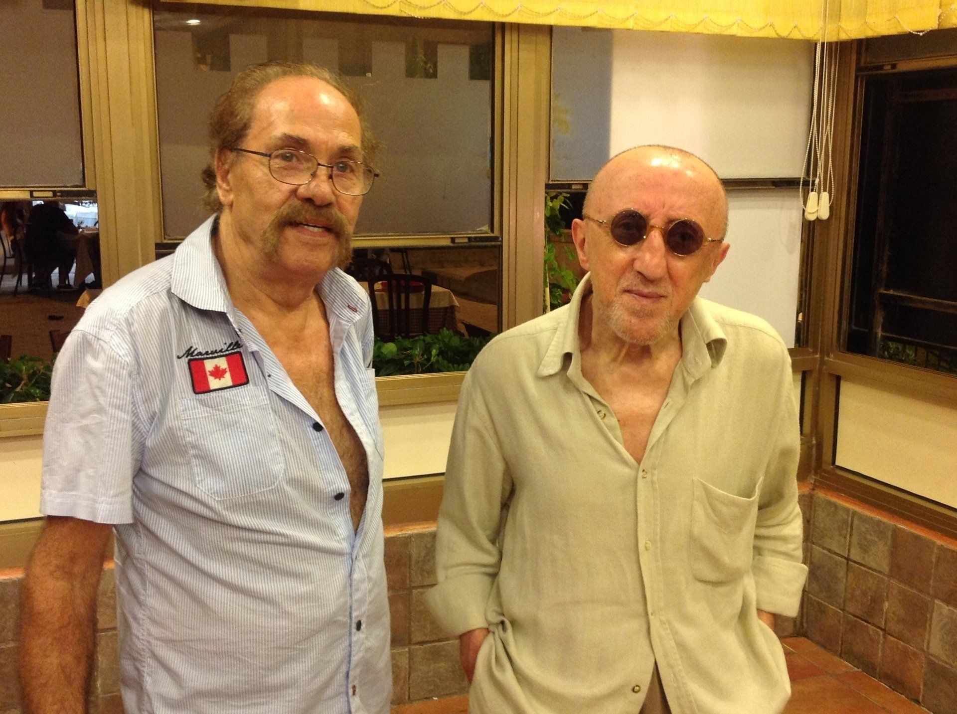 Due uomini in piedi al chiuso, uno con una camicia a righe e l'altro con una camicia verde chiaro e occhiali da sole.