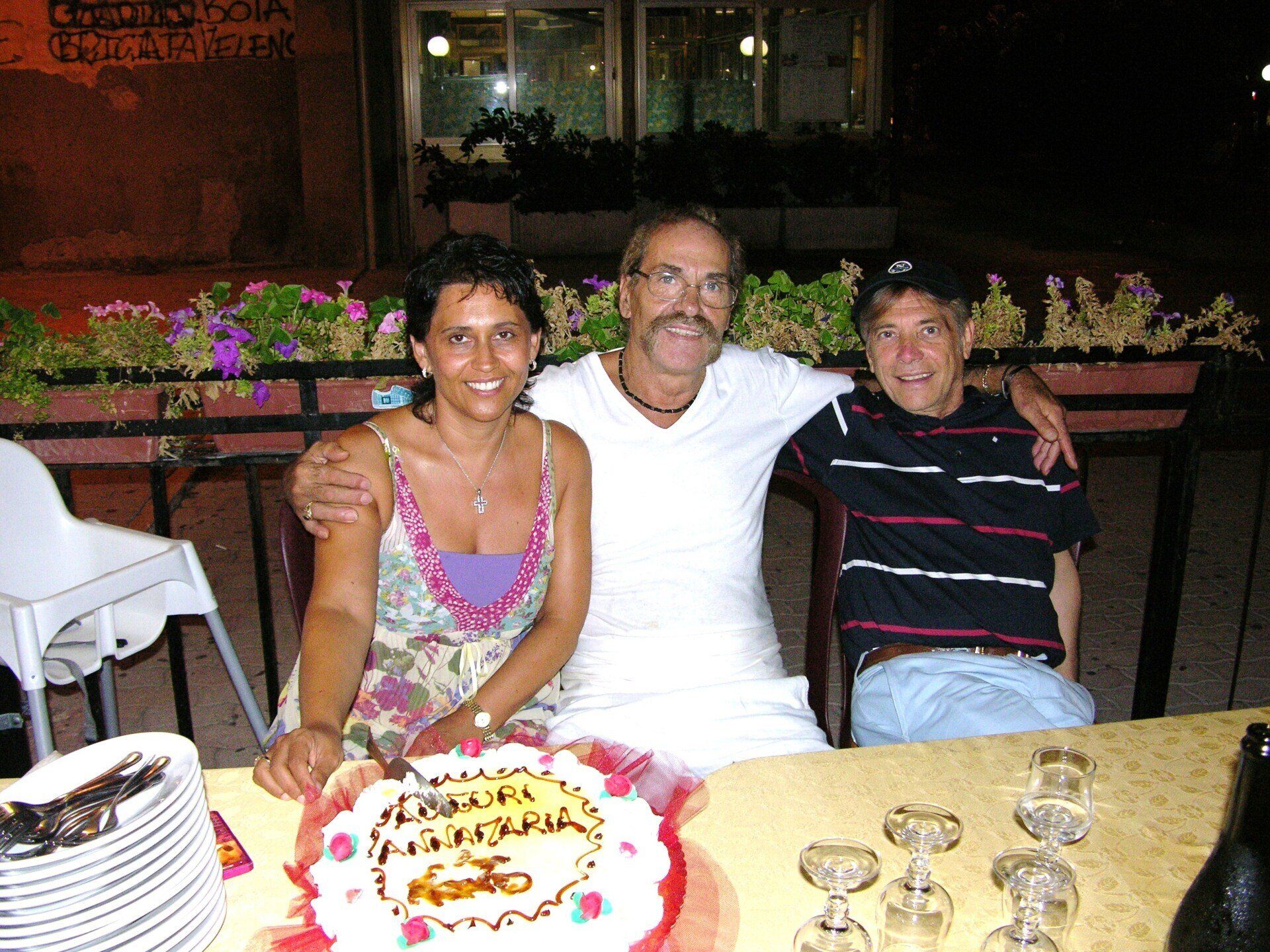 Tre persone sedute a un tavolo all'aperto con una torta decorata e bevande, sorridono durante una festa serale.