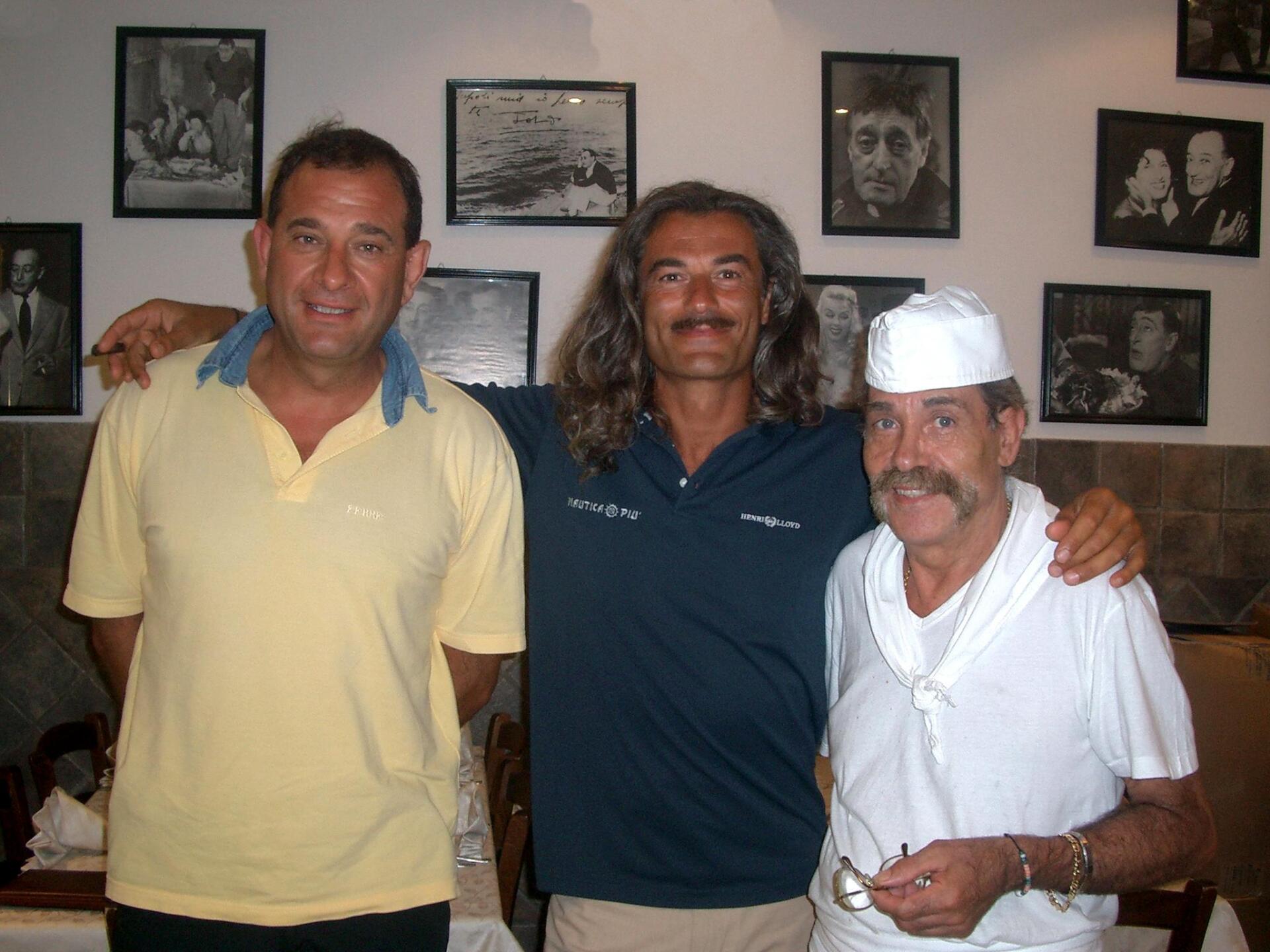 Tre persone in posa in interni, abbracciate, con foto incorniciate appese al muro alle loro spalle.