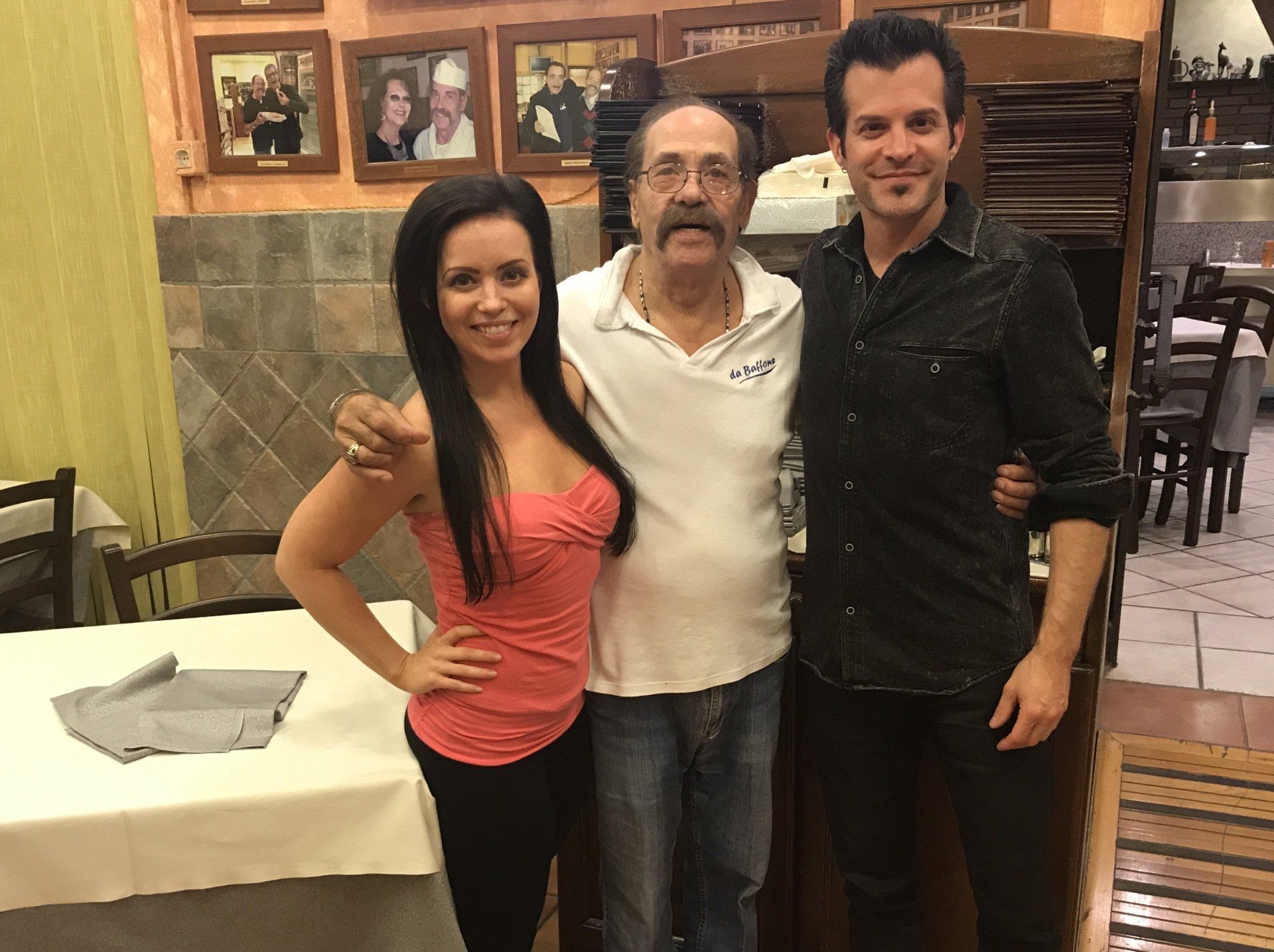 Tre persone in posa insieme in un ristorante, con foto incorniciate e tavoli sullo sfondo.