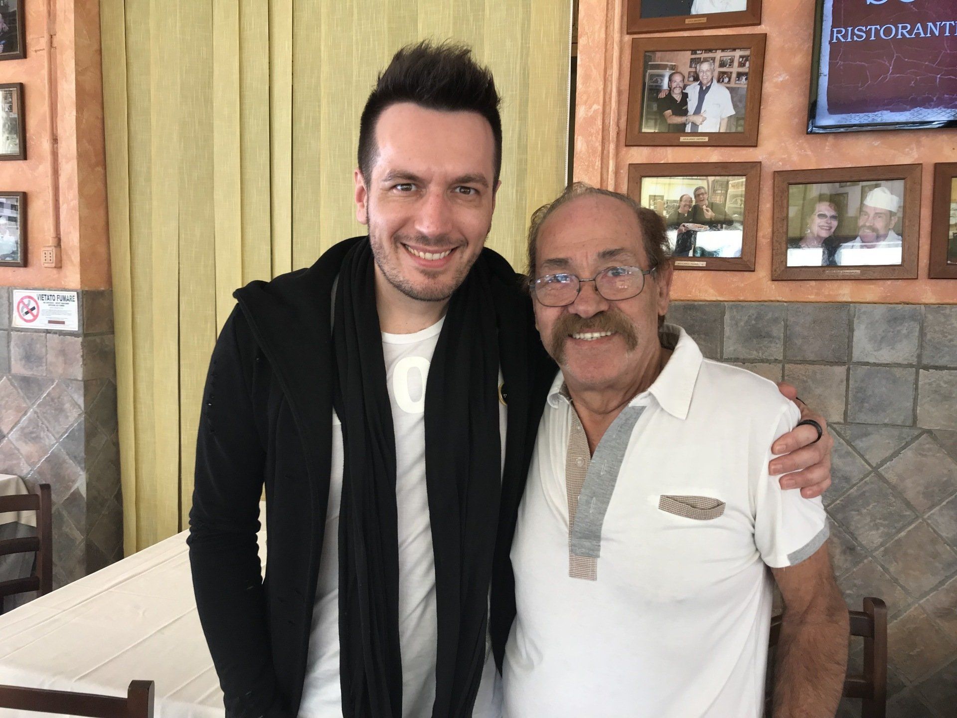 Due uomini sorridenti in piedi uno accanto all'altro in un ambiente chiuso, uno con giacca e sciarpa nere, l'altro con una polo bianca.