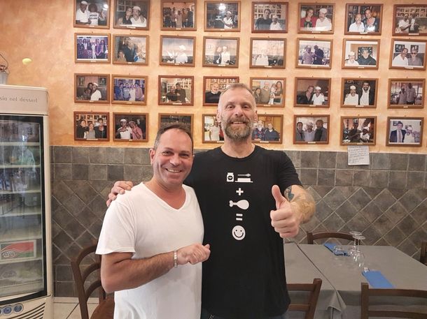 Due uomini sorridenti posano in un ambiente interno davanti a una parete ricoperta di fotografie incorniciate, uno dei quali fa il segno del pollice in su.