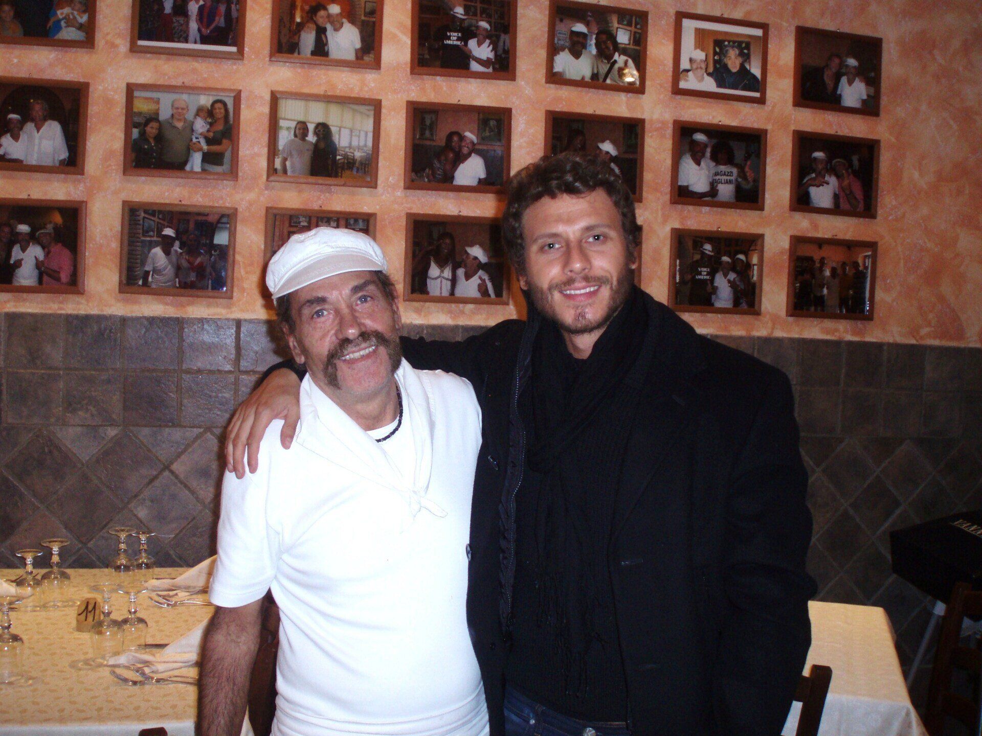 Due uomini in posa insieme in un ristorante, uno con camicia e berretto bianchi, l'altro con giacca nera.