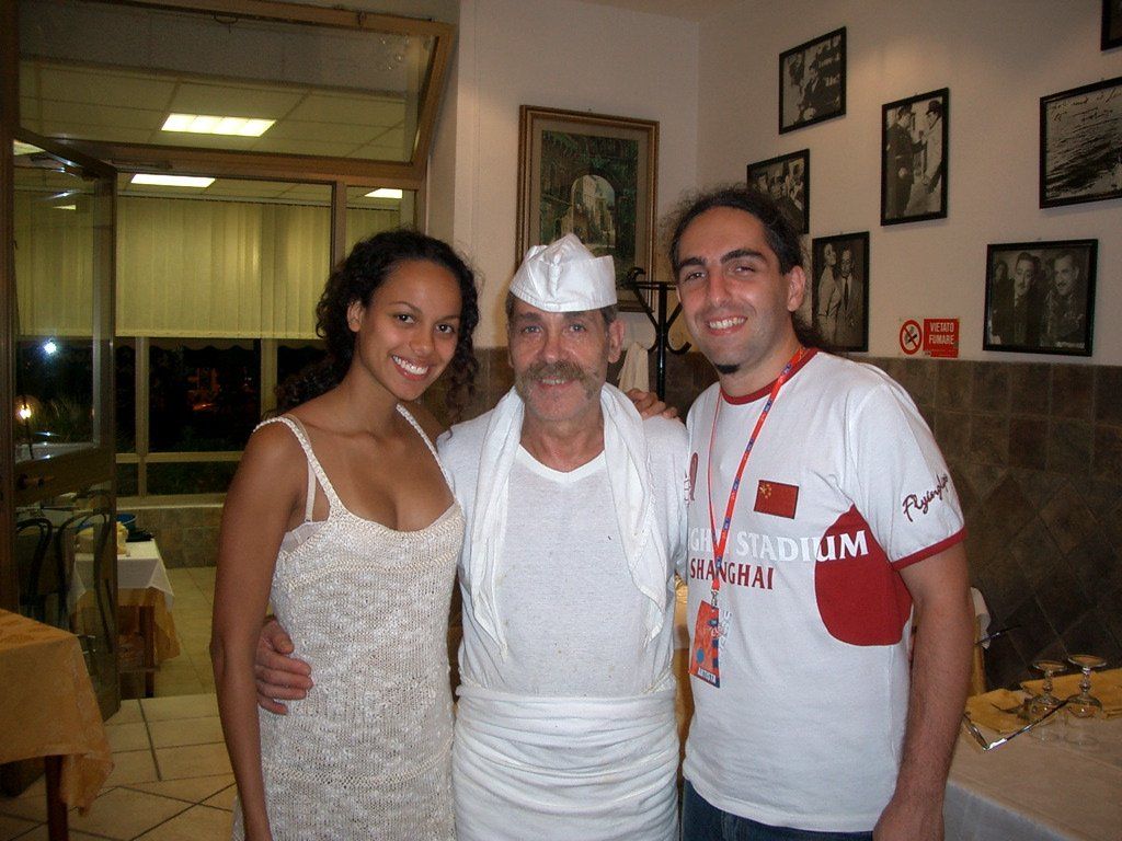 Tre persone in posa insieme in un ambiente interno, una delle quali indossa un cappello da chef e un grembiule.