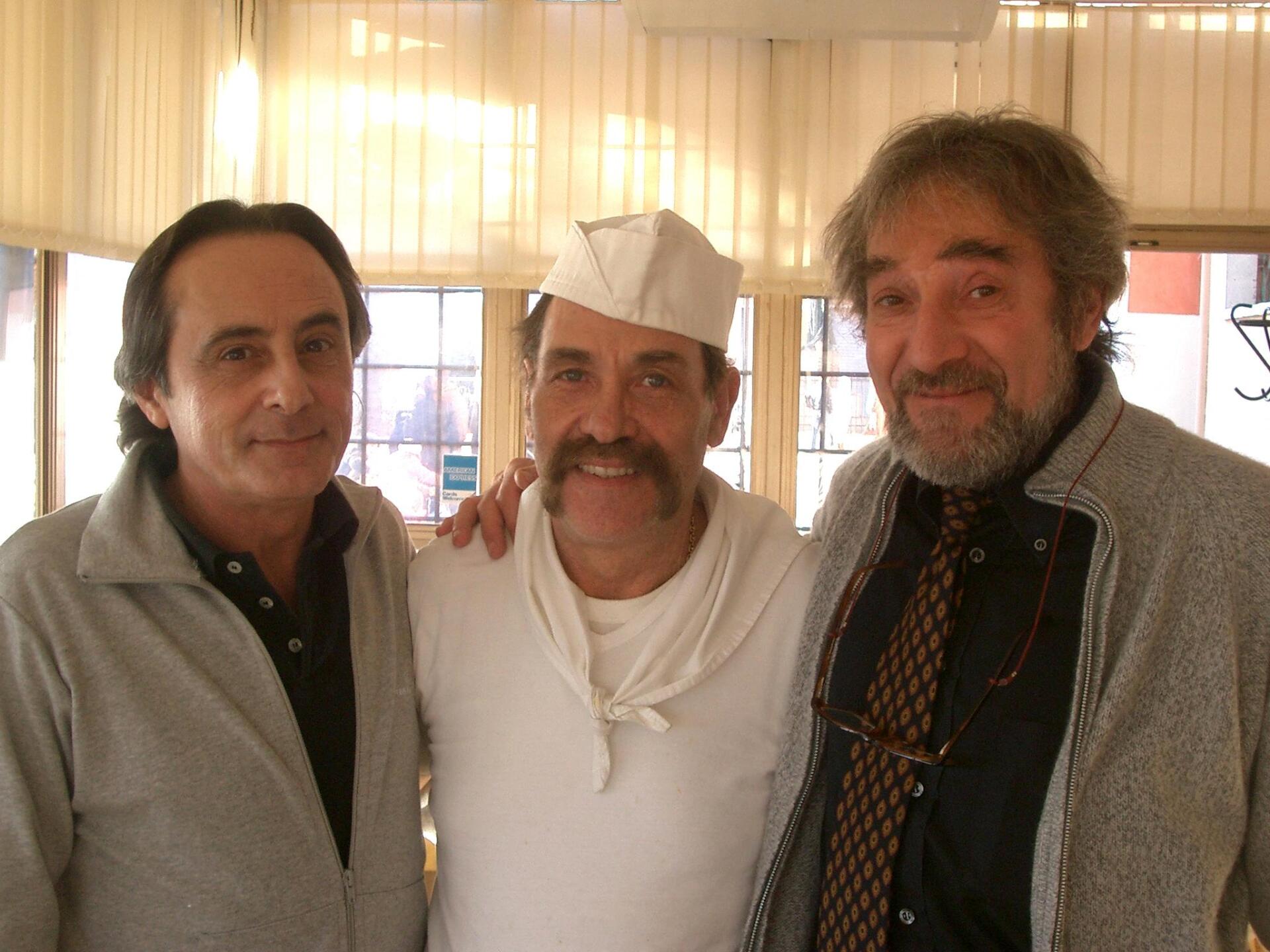 Tre uomini in piedi al chiuso, sorridenti, uno con un cappello di carta bianco e gli altri con delle giacche.