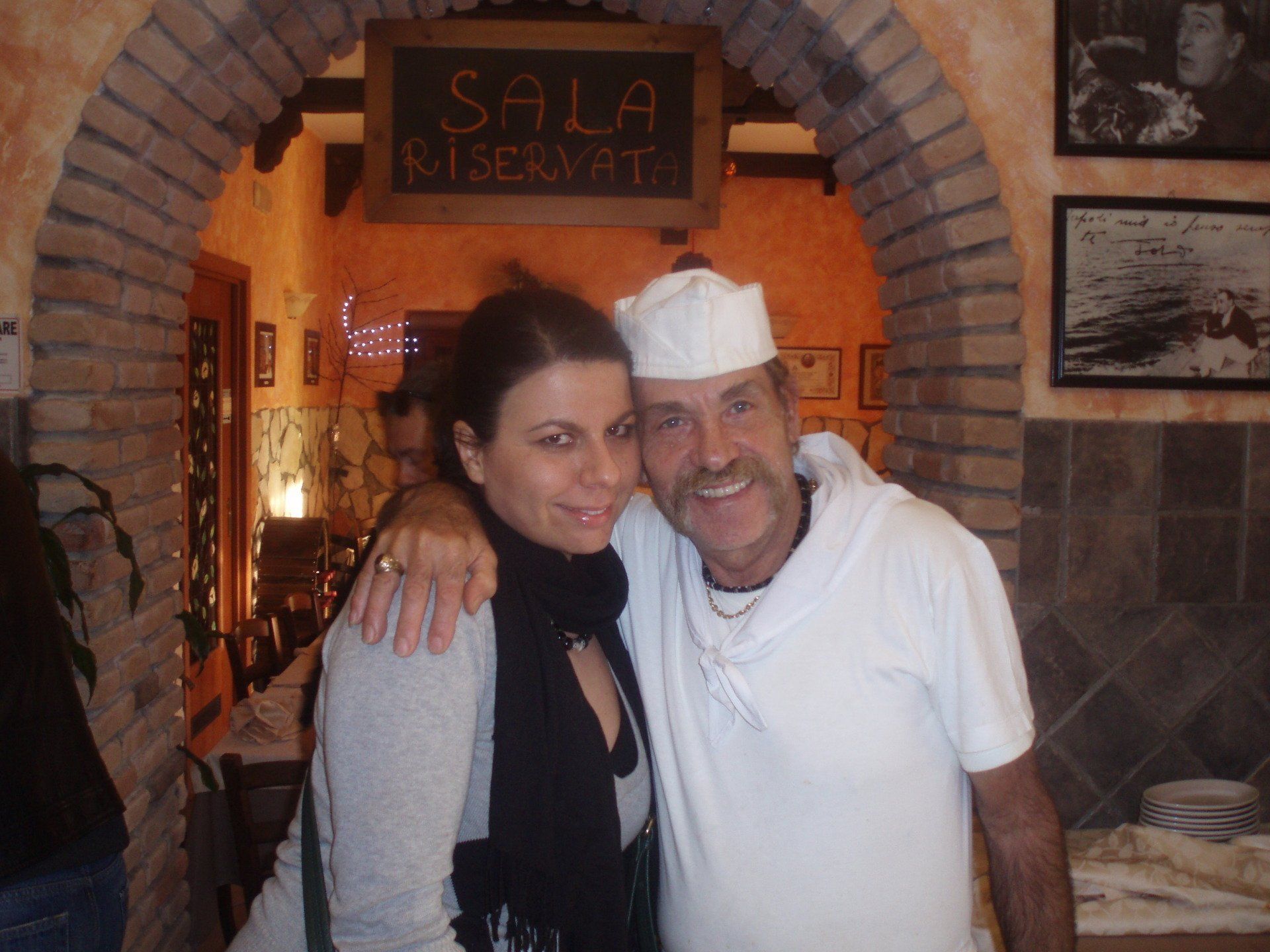 Due persone sorridenti che si abbracciano all'interno dell'ingresso rustico di un ristorante con archi in mattoni.