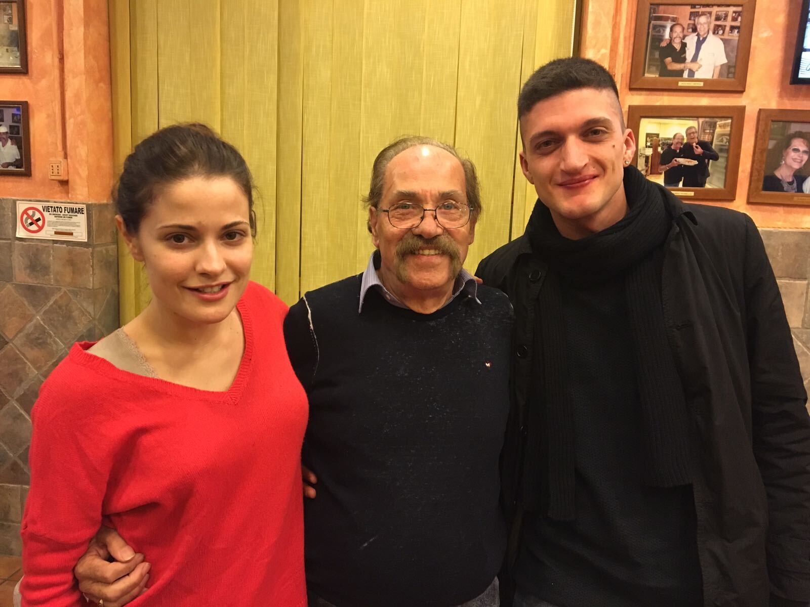 Tre persone in posa in interni, con foto incorniciate appese al muro alle loro spalle.