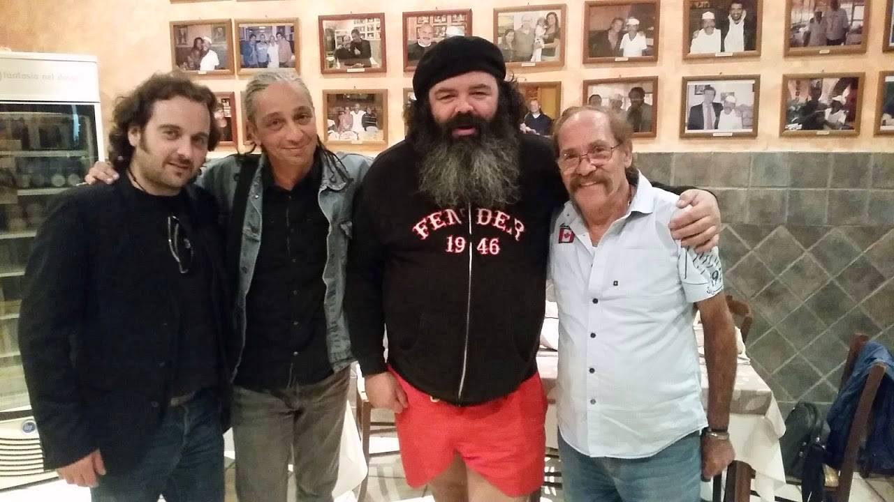 Quattro uomini in posa in interni davanti a una parete ricoperta di foto incorniciate, uno dei quali indossa una felpa nera con cappuccio e pantaloncini rossi.