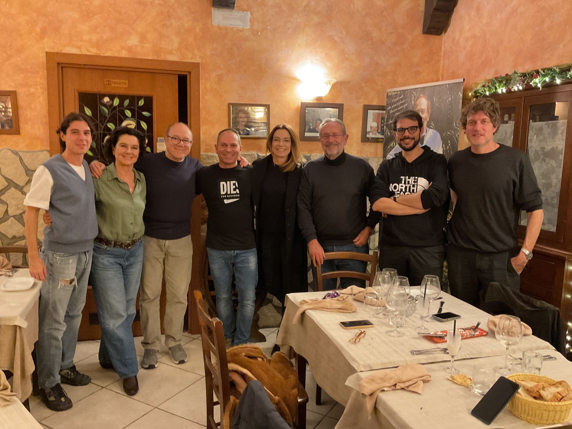 Sei persone posano insieme nella sala da pranzo di un ristorante, illuminata da una luce calda, con un tavolo apparecchiato di fronte a loro.