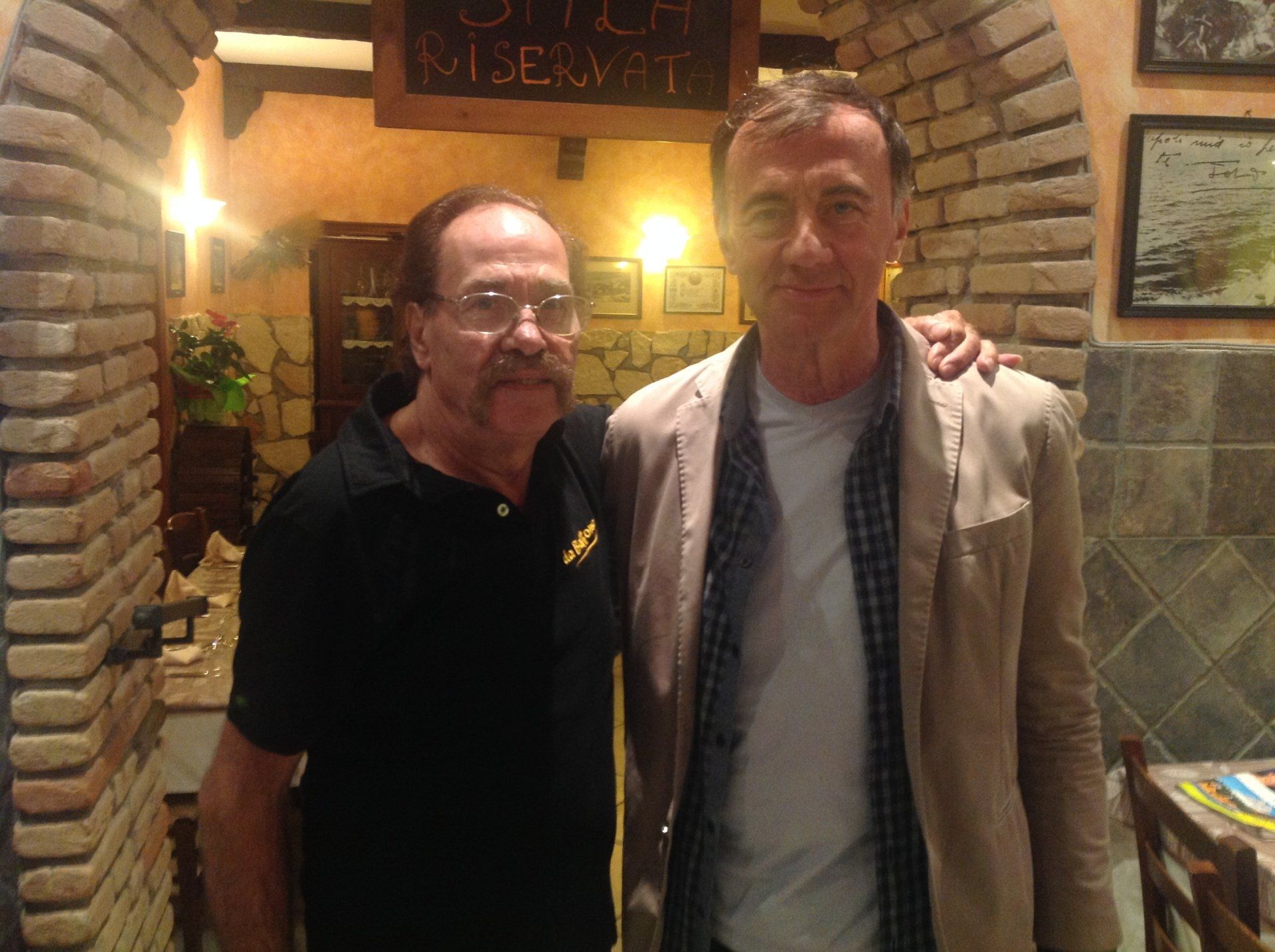 Due uomini posano insieme nell'ingresso di un ristorante in mattoni, illuminato da una luce calda.