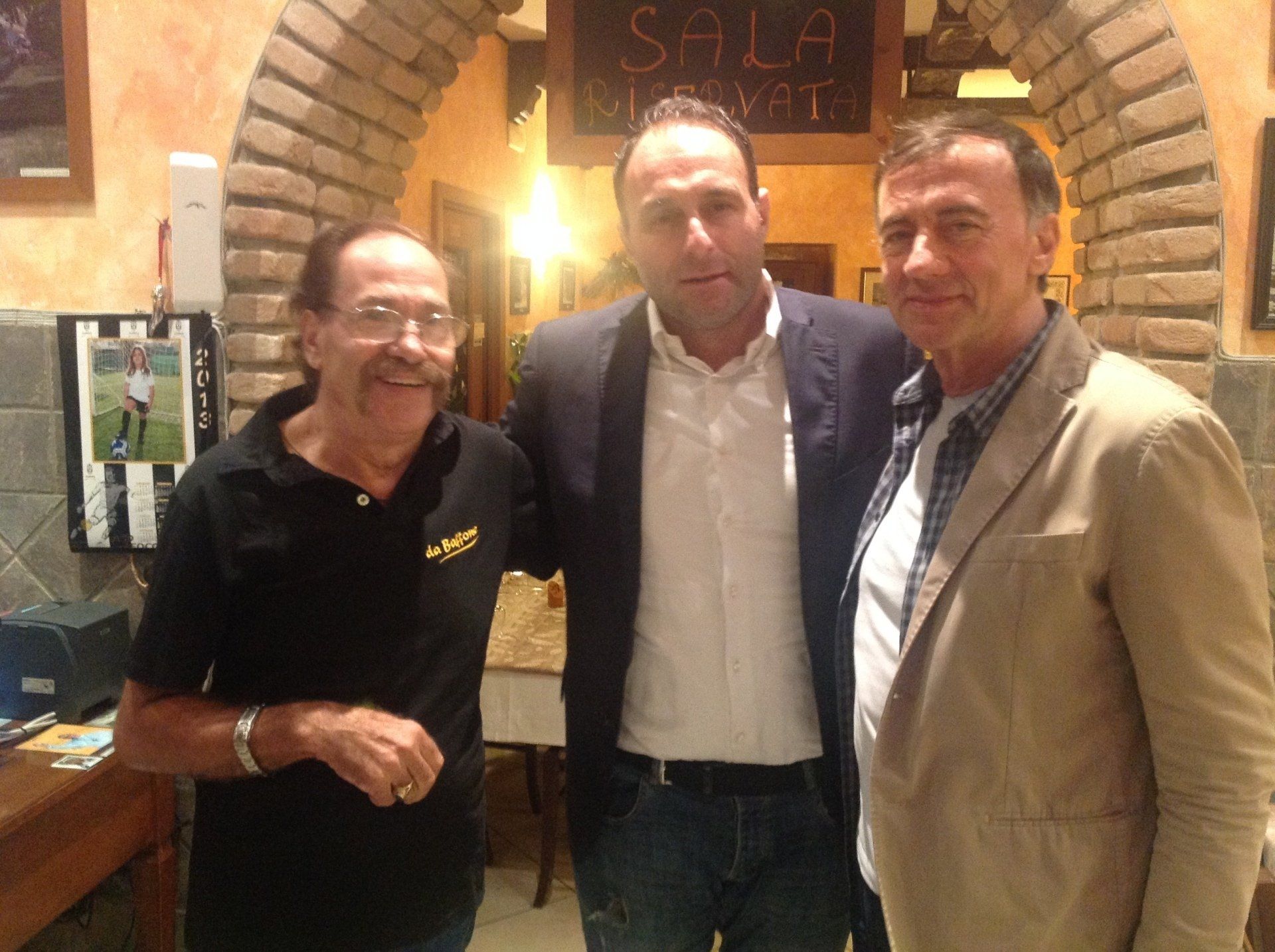 Tre uomini in piedi insieme nell'ingresso di un ristorante illuminato da una luce calda, sorridono alla macchina fotografica.