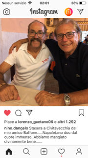 Due uomini sorridenti posano vicini in un ambiente interno, uno con una polo bianca e l'altro con una camicia nera.