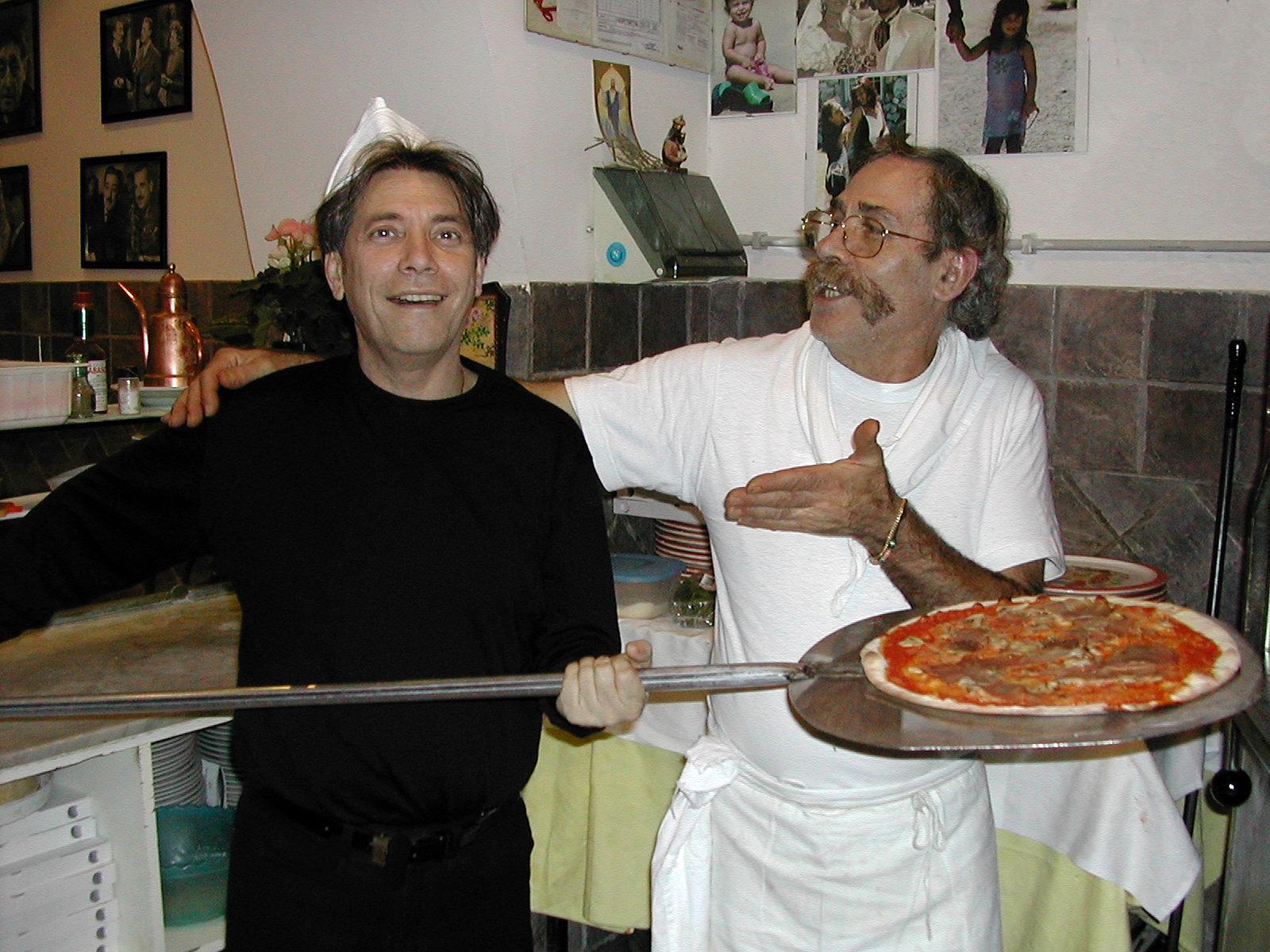 Due persone sorridenti che tengono in mano una pala per pizza con una pizza in cucina