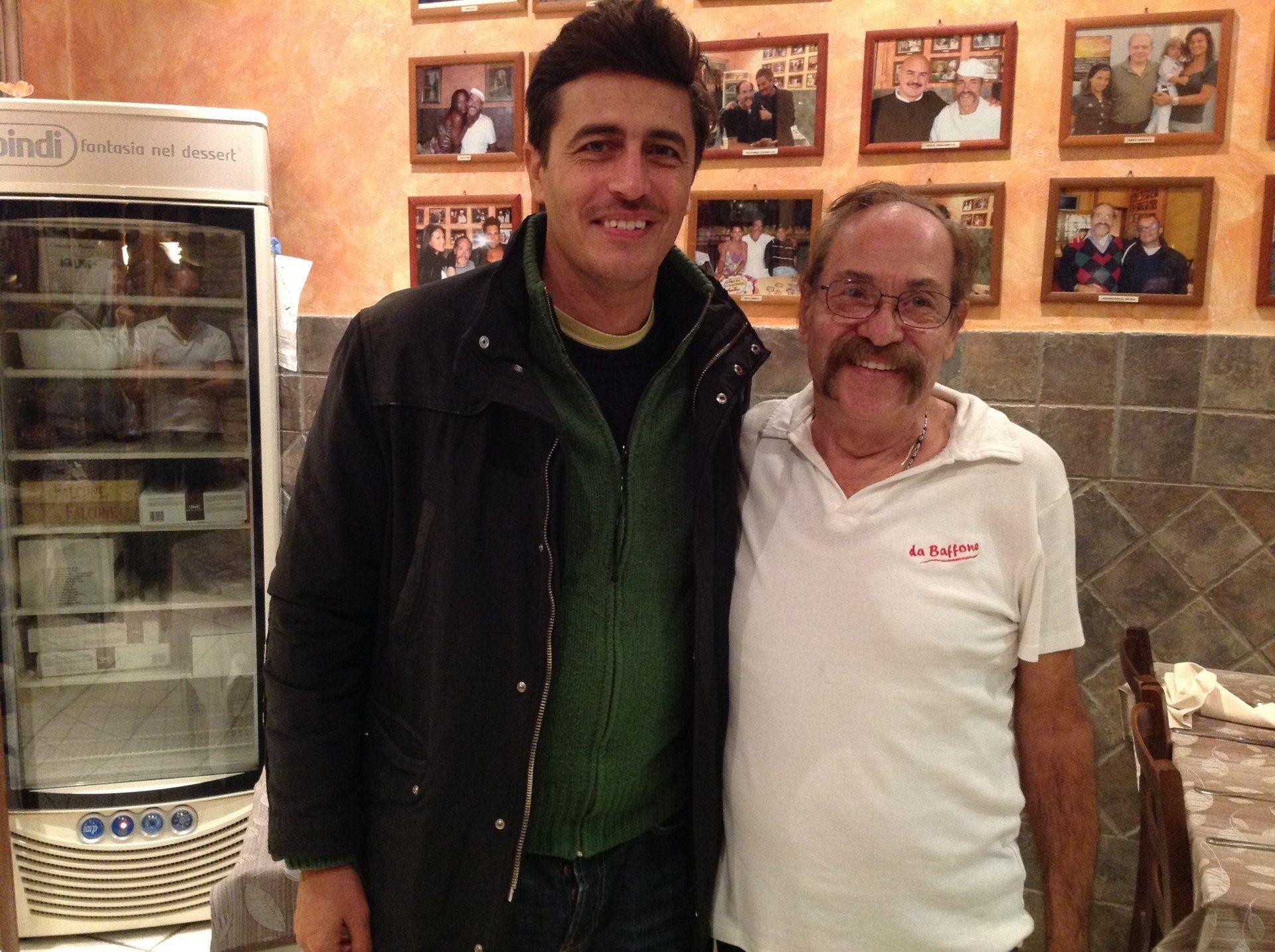 Due uomini in piedi insieme in un ristorante informale, sorridono alla macchina fotografica.
