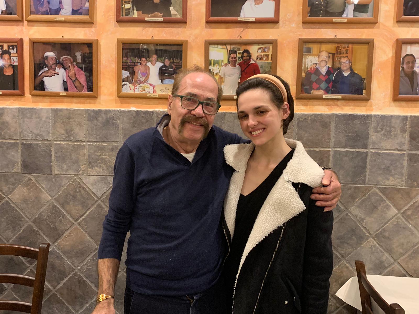 Due persone sorridenti posano insieme in un ristorante, con foto incorniciate appese al muro alle loro spalle.