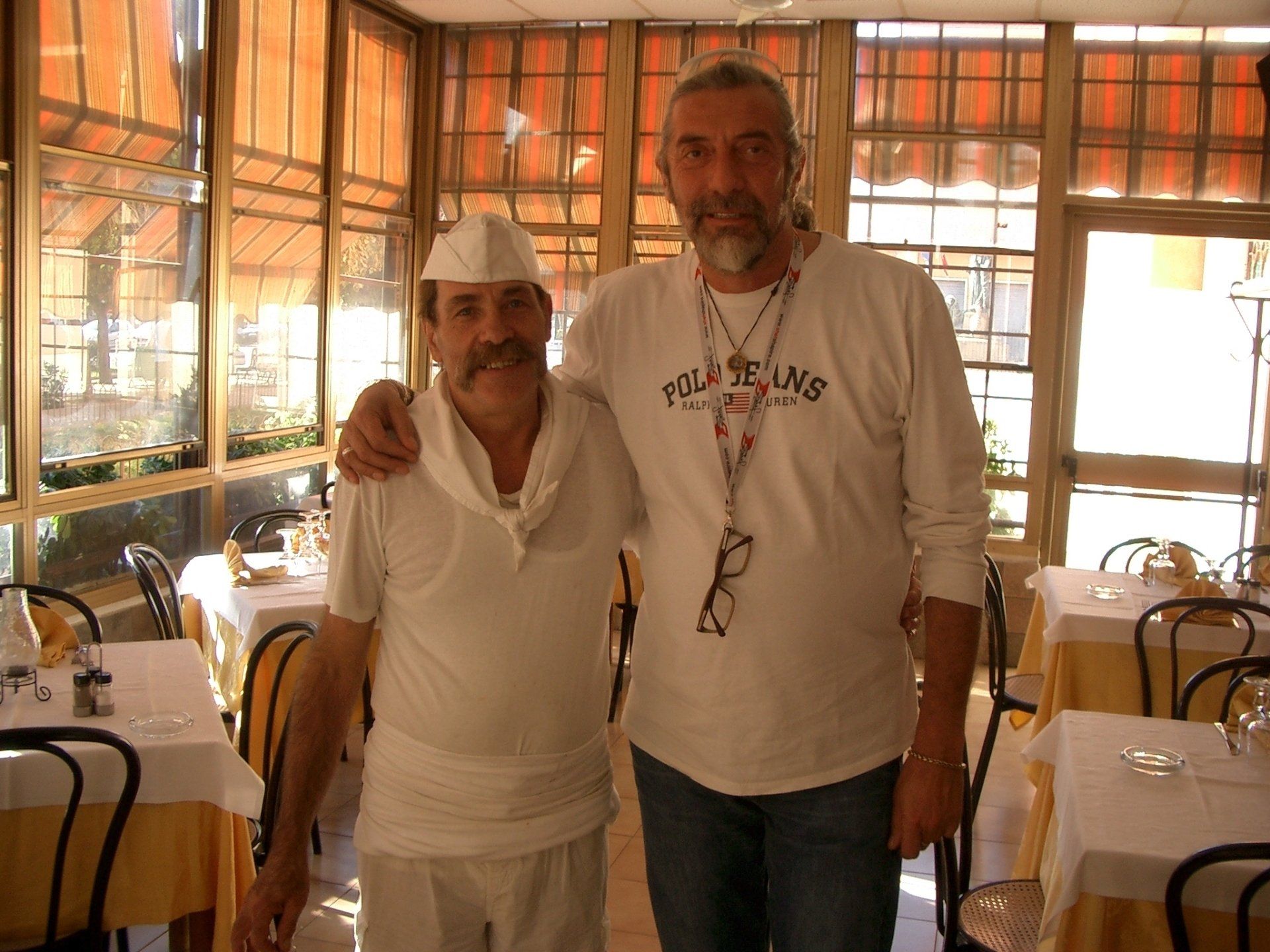 Due persone in piedi insieme in un ristorante luminoso, sorridenti e abbracciate.