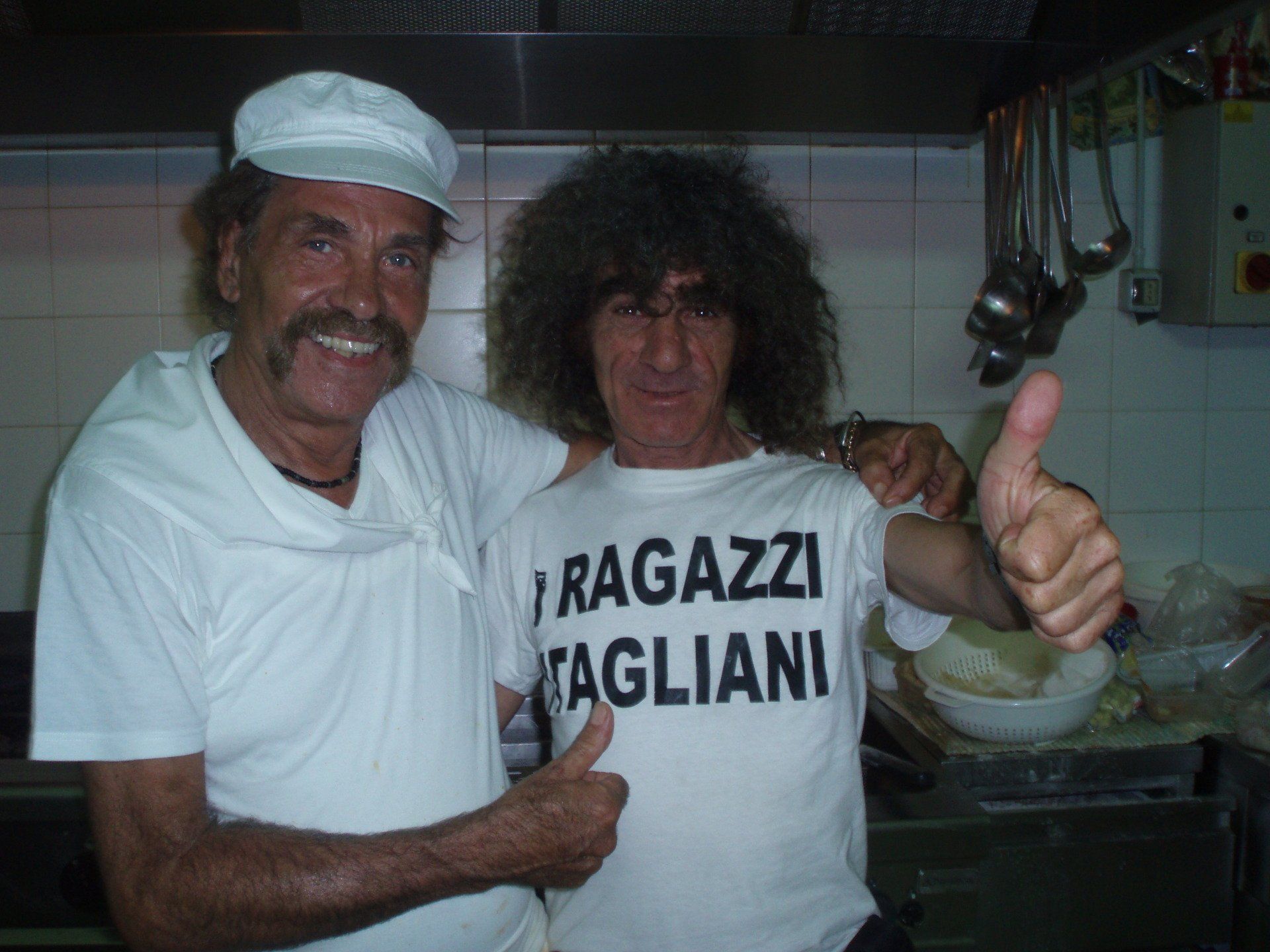 Due persone in cucina, sorridenti e con il pollice alzato, indossano camicie bianche e cappelli da chef.
