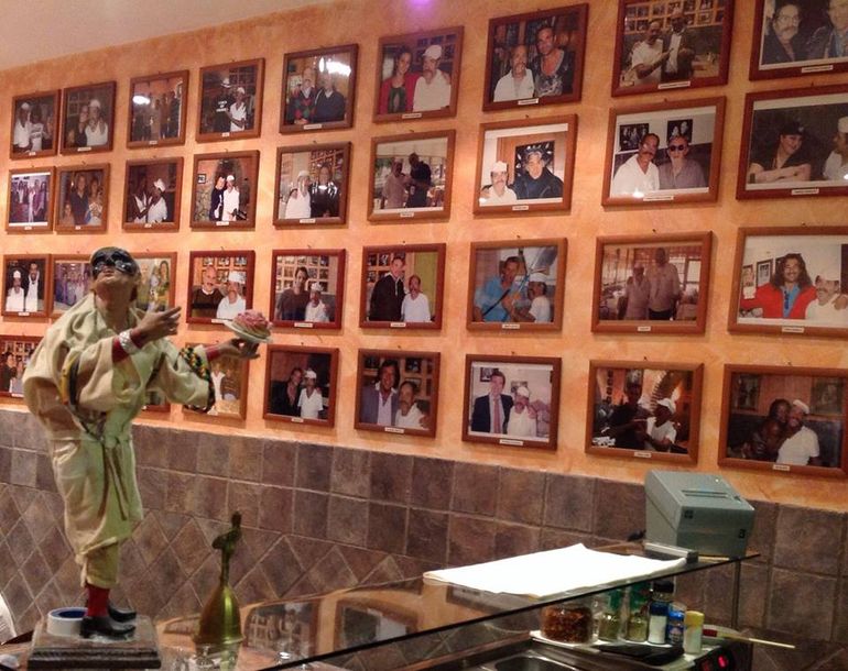 Un manichino vestito di verde posa davanti a una parete di foto incorniciate in una stanza dai toni caldi.
