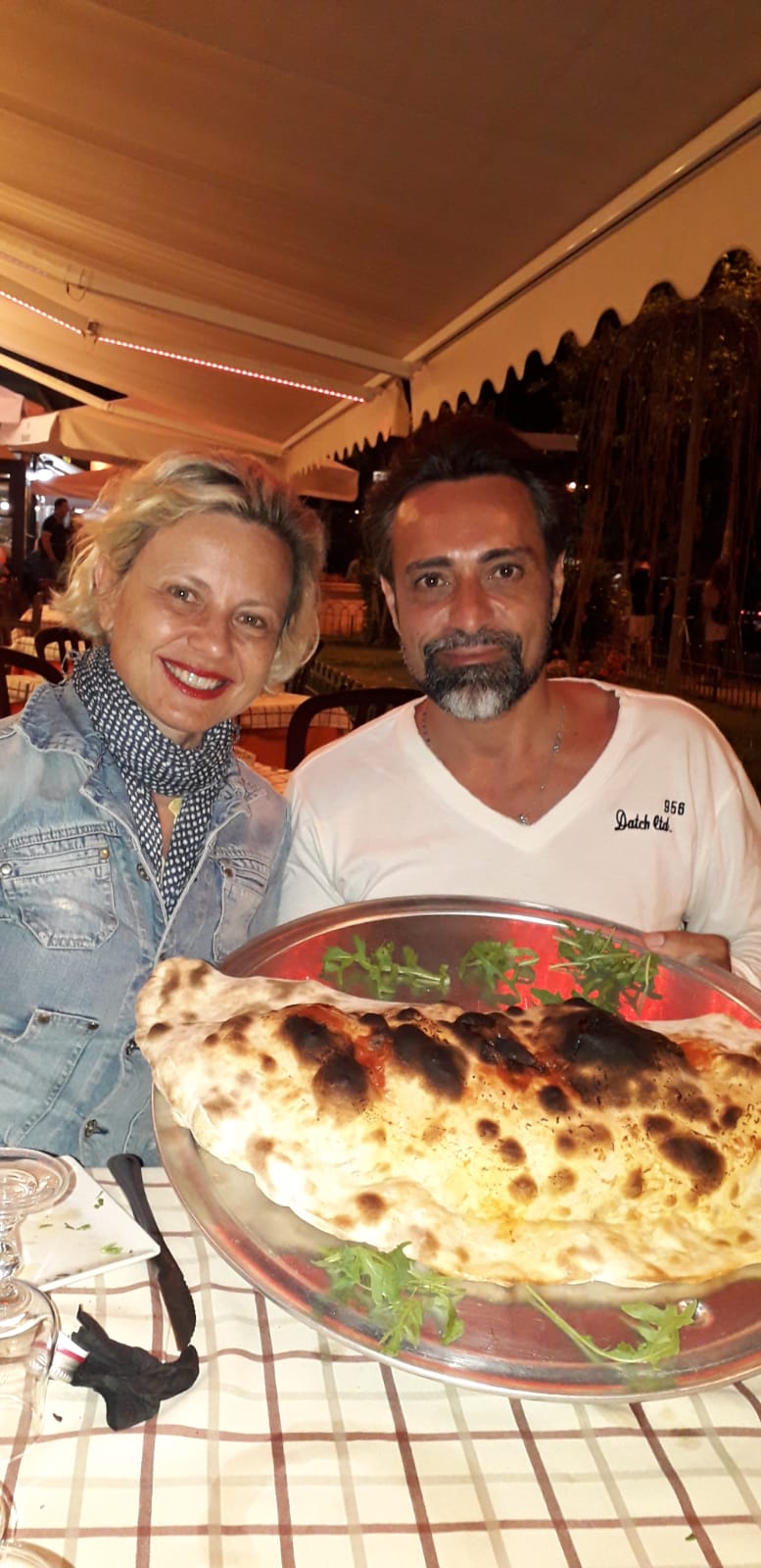 Due persone sorridenti sedute a un tavolo con una grande pietanza al forno, in un accogliente ristorante al chiuso.