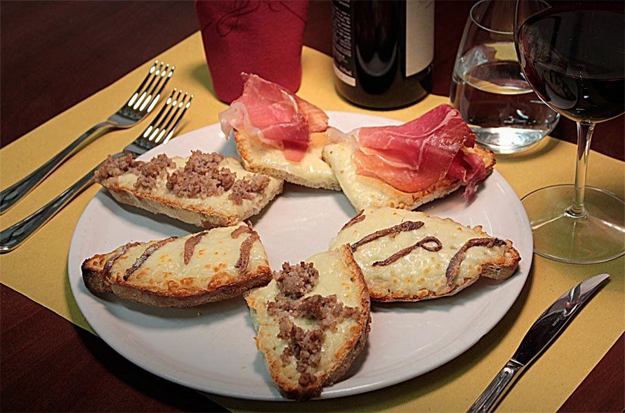Piatto di fette di pane tostato con formaggio e prosciutto su un tavolo di ristorante con bicchieri da vino.