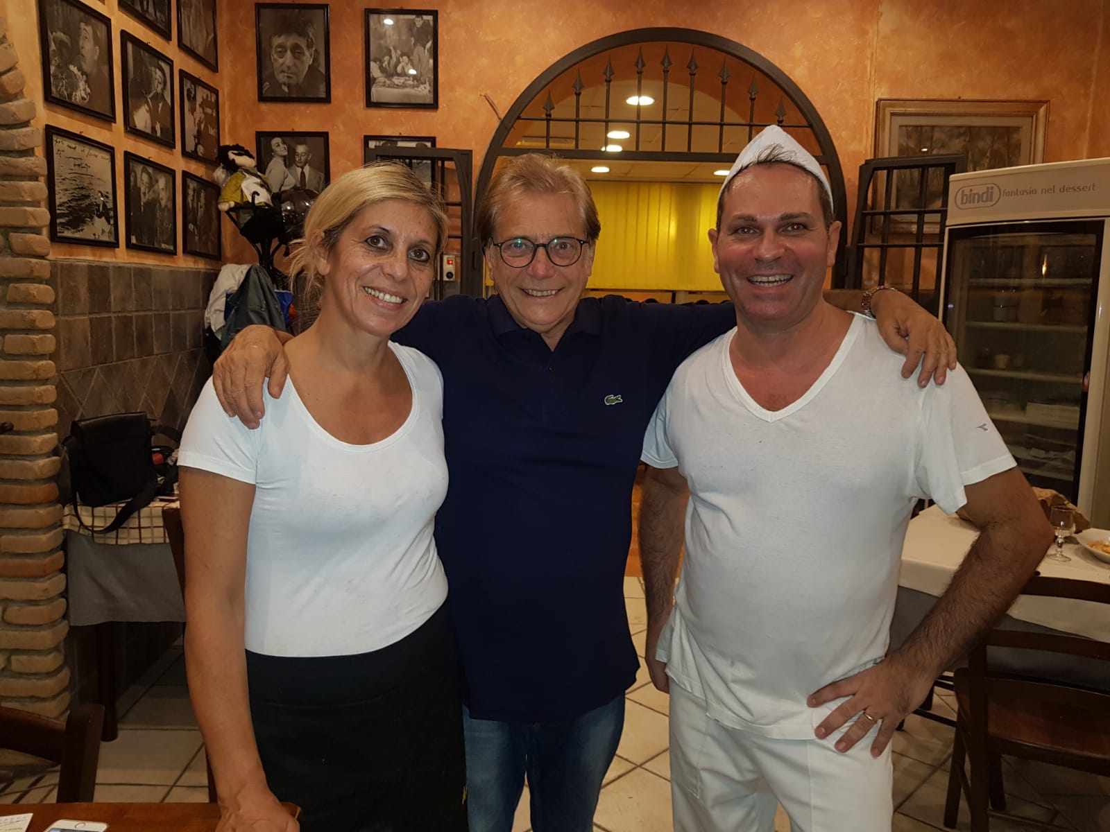 Tre persone sorridenti che posano insieme in un accogliente ristorante con pareti in mattoni e foto incorniciate.